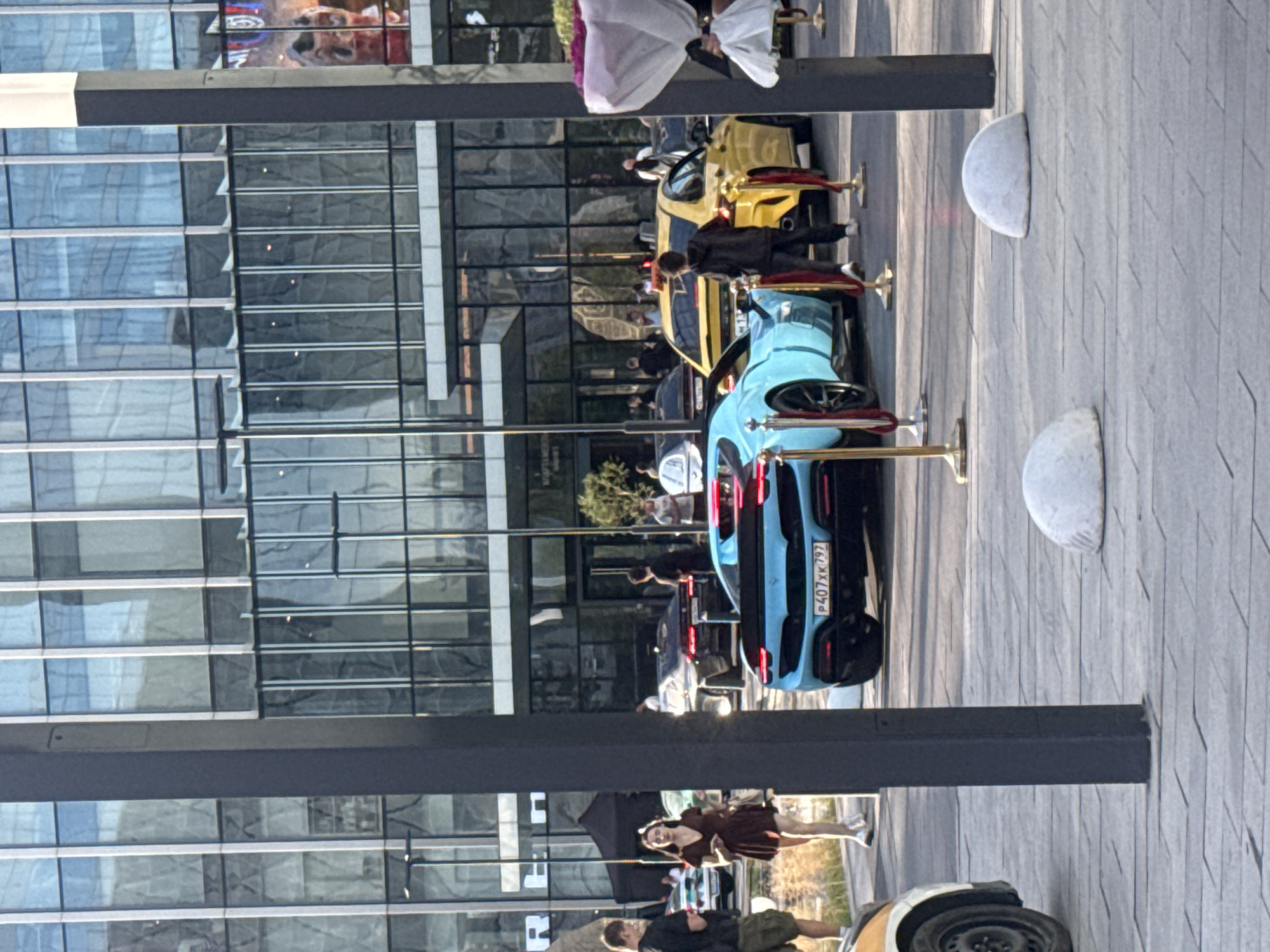 In Khimki, Russia, a street-level view captures a modern urban setting with a large glass-fronted building. In the foreground, a bright blue sports car with license plate P407XK797 is parked, with red velvet ropes and gold stanchions delineating its area. To its right, a yellow sedan is also visible. Several individuals are present, some standing near the cars and others walking in the background. A woman in a burgundy dress is walking towards the left side of the frame, while two men stand near a white car on the far left. The environment appears to be an outdoor plaza or walkway in front of commercial or office buildings, with a clear sky.
