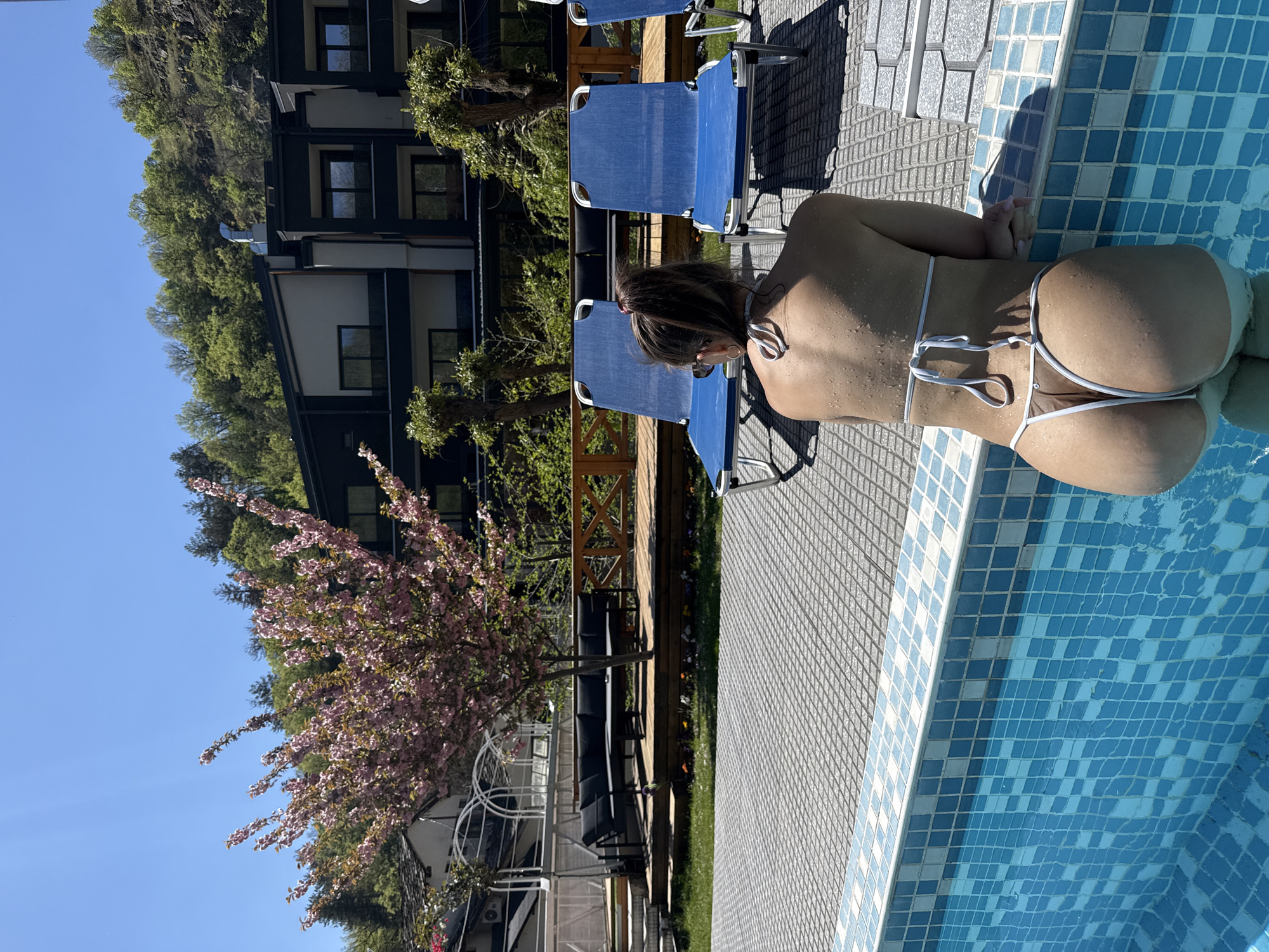 A woman in a white bikini is sitting on the edge of a swimming pool. The pool has blue and white mosaic tiles. The surrounding area is paved with gray tiles. There are blue lounge chairs nearby. In the background, there is a building, trees, and a flowering tree. The location is Marchevo, Bulgaria.