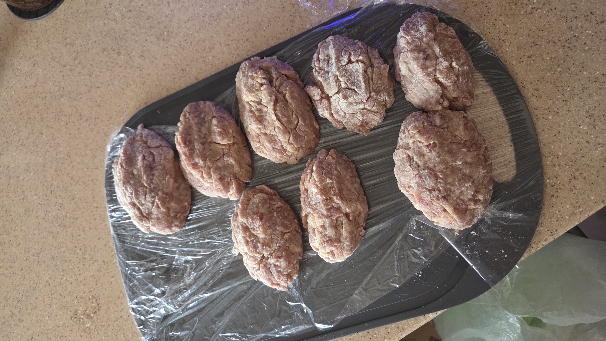 The image displays eight raw, oval-shaped meat patties, possibly cutlets or koftas, meticulously arranged on a dark grey, oval-shaped cutting board or tray. The patties, varying slightly in size and shape, are made from ground meat, exhibiting a pale pinkish-brown color and a somewhat textured surface. Some of them appear to have a light dusting, possibly of flour or breadcrumbs, contributing to their texture.
The dark grey board is partially covered with clear, crinkled plastic wrap, which reflects some ambient light, suggesting the patties are either being prepared for immediate cooking or for storage. The board rests on a light brown, speckled countertop, which forms the background and a significant portion of the setting. In the bottom right corner of the frame, a blurry, translucent light green object, possibly a plastic bag, is faintly visible.
The scene captures a moment of food preparation in what appears to be an indoor kitchen setting. The lighting suggests it is daytime. There are no people visible, and no specific activities beyond the static arrangement of the patties are depicted. No text is visible within the image. The described context places this scene in Chaikovsky, Russia.