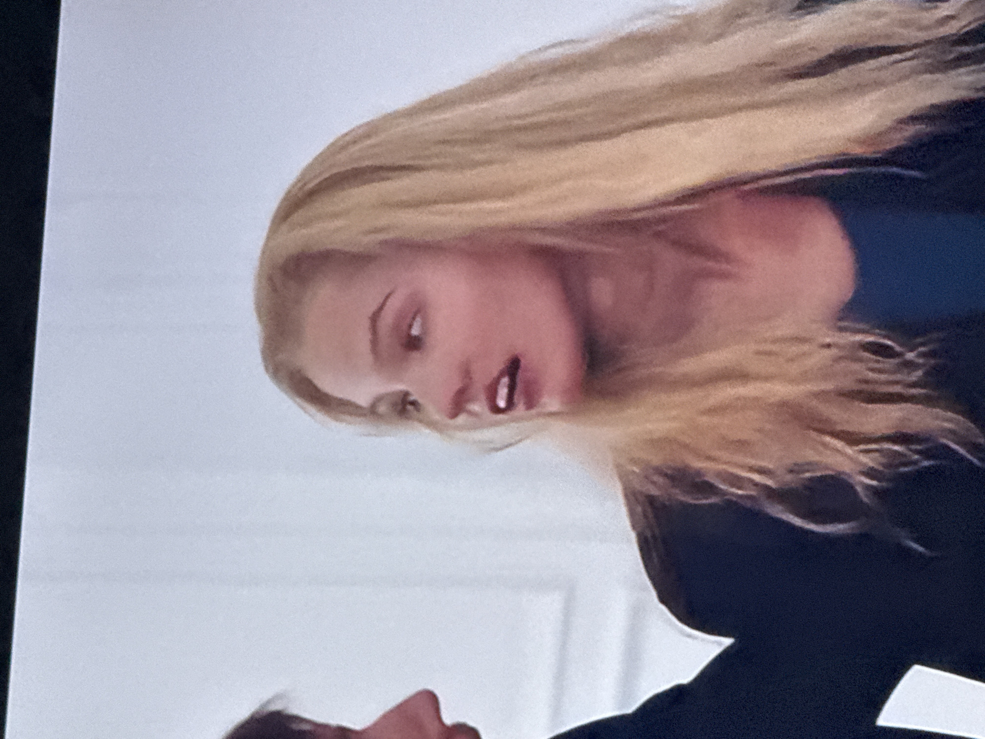 A woman with long blonde hair and a blue shirt is facing downward. Part of another person's face is visible below her. The background is a white wall, possibly with a doorframe. The image location is not discernible from the provided information.