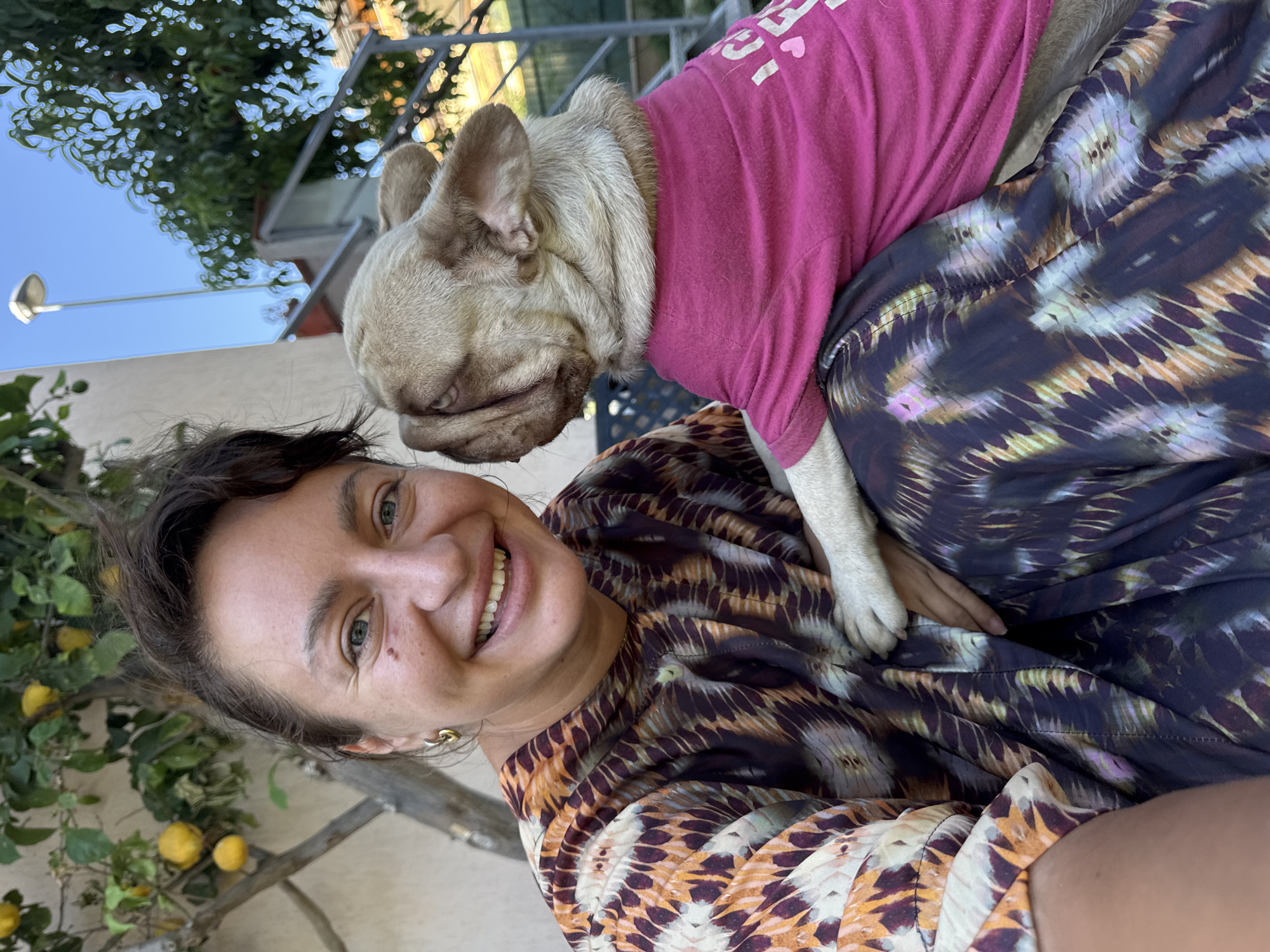 The image shows a close-up, outdoor, eye-level shot of a woman and a French bulldog. The woman is smiling and looking at the camera. She has dark hair pulled back and is wearing a colorful patterned top. The French bulldog, fawn-colored, is wearing a bright pink t-shirt and is nuzzling the woman's face. They are seated outdoors, with a lemon tree visible behind the woman and a blurred background of greenery, a wall, and a railing, suggesting a residential courtyard in San Benedetto del Tronto, Italy. The lighting indicates it is daytime.