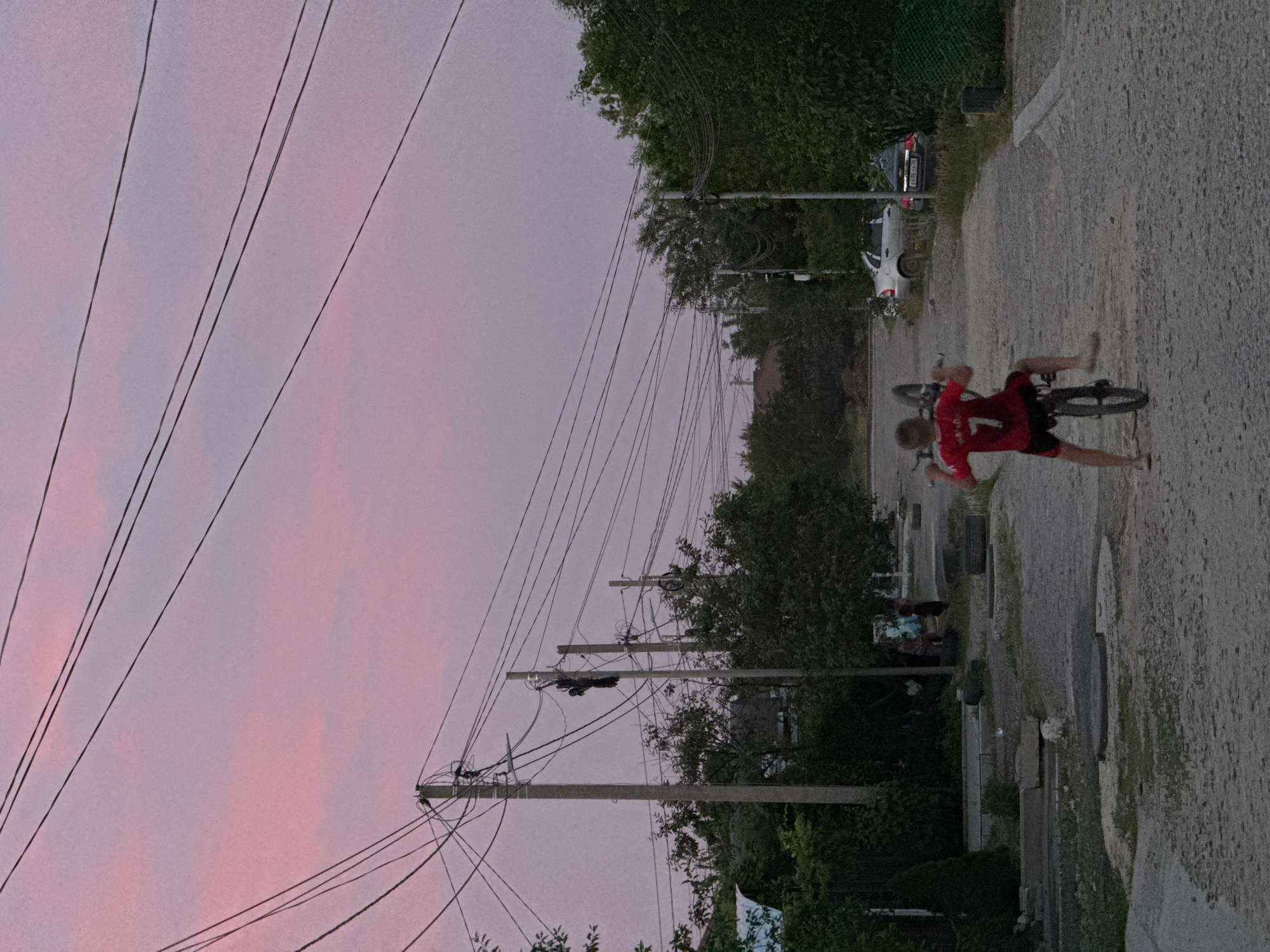 A young boy in a red shirt with the number 7 is performing a wheelie on a bicycle on a gravel road. He is facing away from the camera, toward the right side of the frame. His shirt has a white number 7 on the back. He is wearing black shorts and appears to be barefoot or wearing very light sandals. The bicycle's front wheel is raised in the air. The road is lined with trees and houses. In the background, there are power lines stretching across the sky. The sky is a soft pink and purple, suggesting it is either sunrise or sunset. Several cars are parked along the side of the road. The environment appears to be a residential area in Mikhaylovsk, Russia.