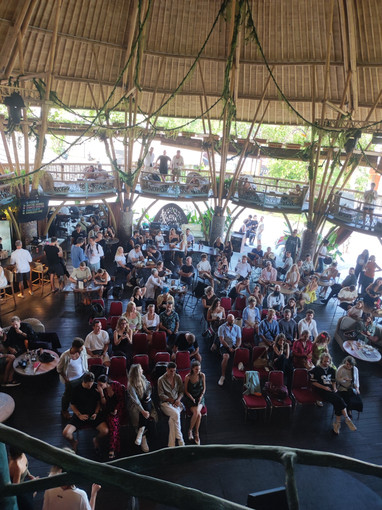 The image shows a large, open-air structure, possibly a restaurant or event space, in an unknown city in Indonesia. The building has a high, bamboo-supported roof with hanging greenery. Multiple levels are visible, with people on each level. The primary level features rows of red chairs arranged as if for a presentation or performance. People are seated in these chairs and others are sitting at tables and in lounging areas scattered around the space. On the upper levels, people are standing along the railing or seated at tables. Patrons are engaged in conversations and activities associated with dining and entertainment.