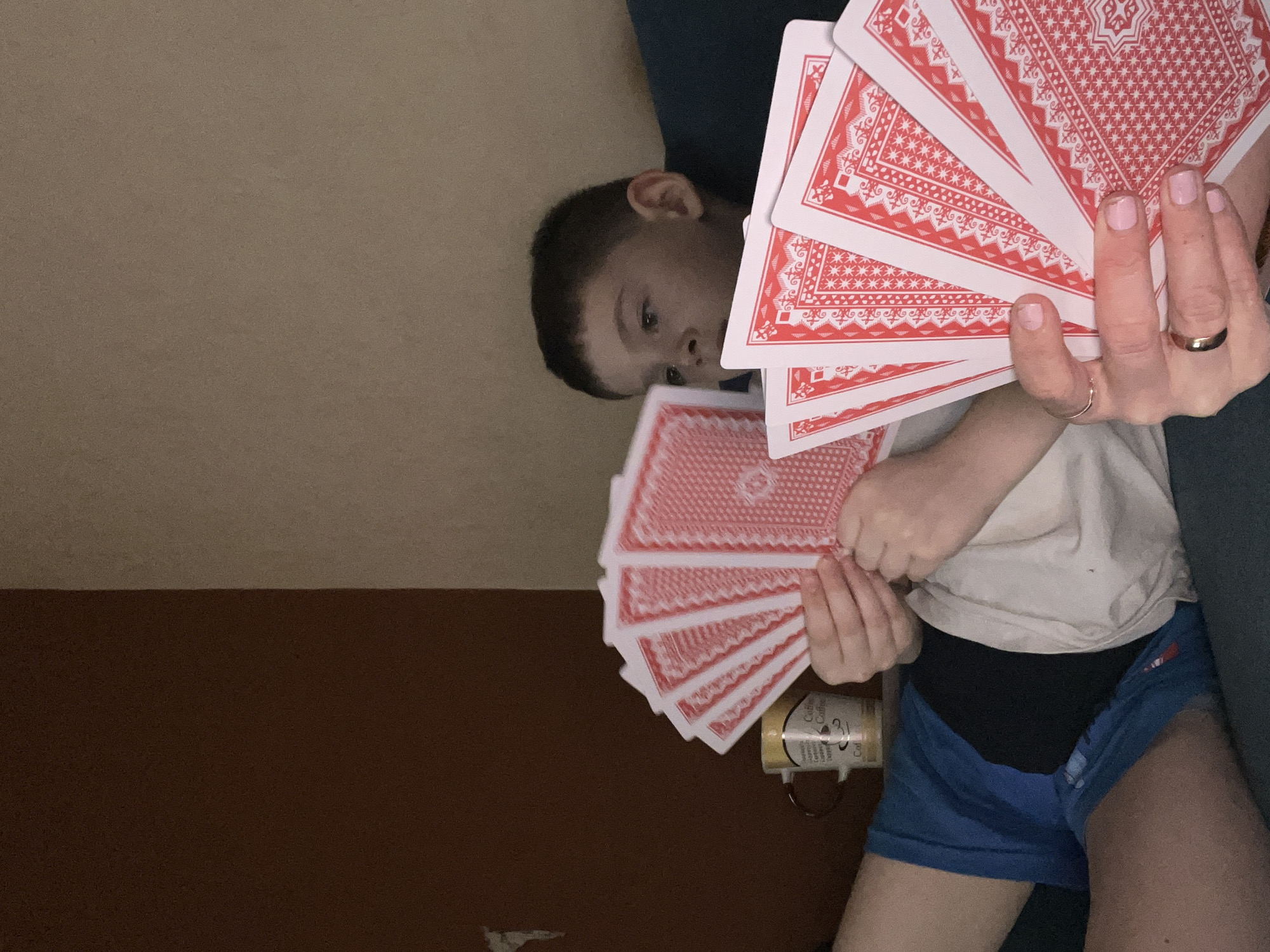 The image depicts an indoor scene featuring an adult and a young boy, both holding fanned playing cards. The image is rotated 90 degrees clockwise.
In the upper right, an adult's hand with light pink manicured nails and two gold rings holds a fan of red-backed playing cards. Below this, a young boy with short, dark hair is peeking from behind another fan of red-backed playing cards that he is holding. He is wearing a light-colored t-shirt over blue shorts, with a dark undershirt visible beneath his t-shirt. His eyes are directed towards the viewer, conveying a sense of curiosity or engagement in a game.
The setting appears to be a comfortable indoor space. A dark green or blue fabric, likely a couch or blanket, is visible behind the adult's hand and the boy's head. The background consists of a plain brown wall, with a lighter section above it. Next to the boy, a brown mug or container with a handle is partially visible. The word "Coffee" is discernable on its side, along with other unreadable text. The lighting is dim, suggesting evening or an ambient indoor environment. There are no specific cues within the image to indicate the location as Chaikovsky, Russia.