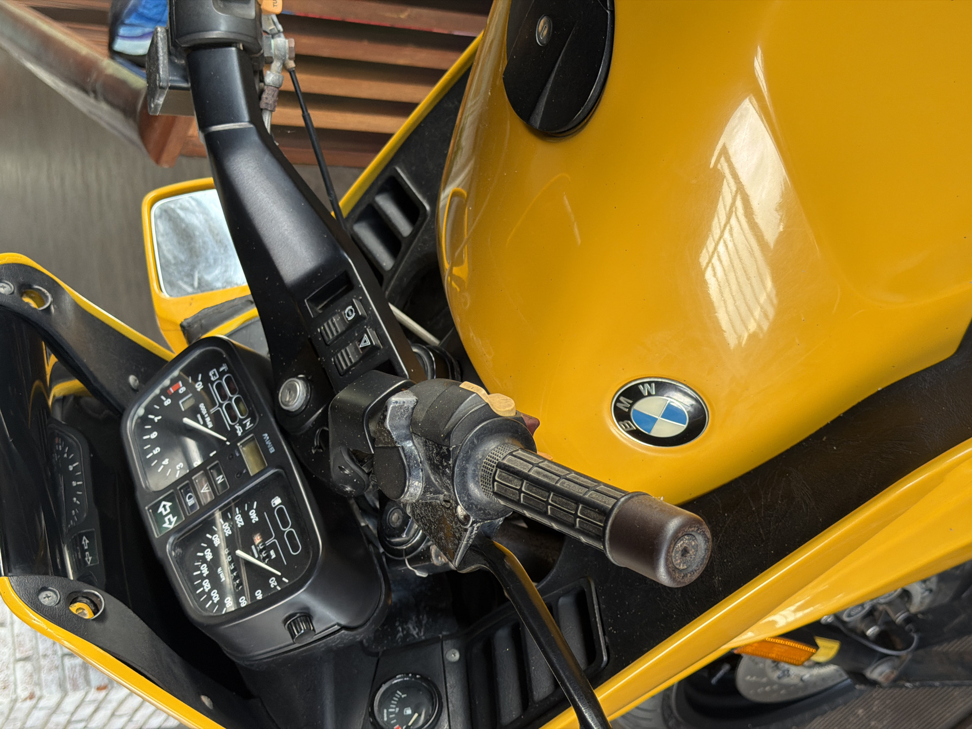 The image shows a close-up, eye-level view of a yellow BMW motorcycle's handlebars and front section. The motorcycle is parked outdoors, with a blurred background of wooden slats and a hint of a brick wall. The handlebars are black and feature a textured grip on the right side. To the left of the handlebars, a detailed instrument cluster is visible, displaying a speedometer, tachometer, and various indicator lights. The BMW logo is prominently featured on the yellow fuel tank. The overall scene suggests a still, observational shot of the motorcycle. The location is specified as Gerokgak, Indonesia.