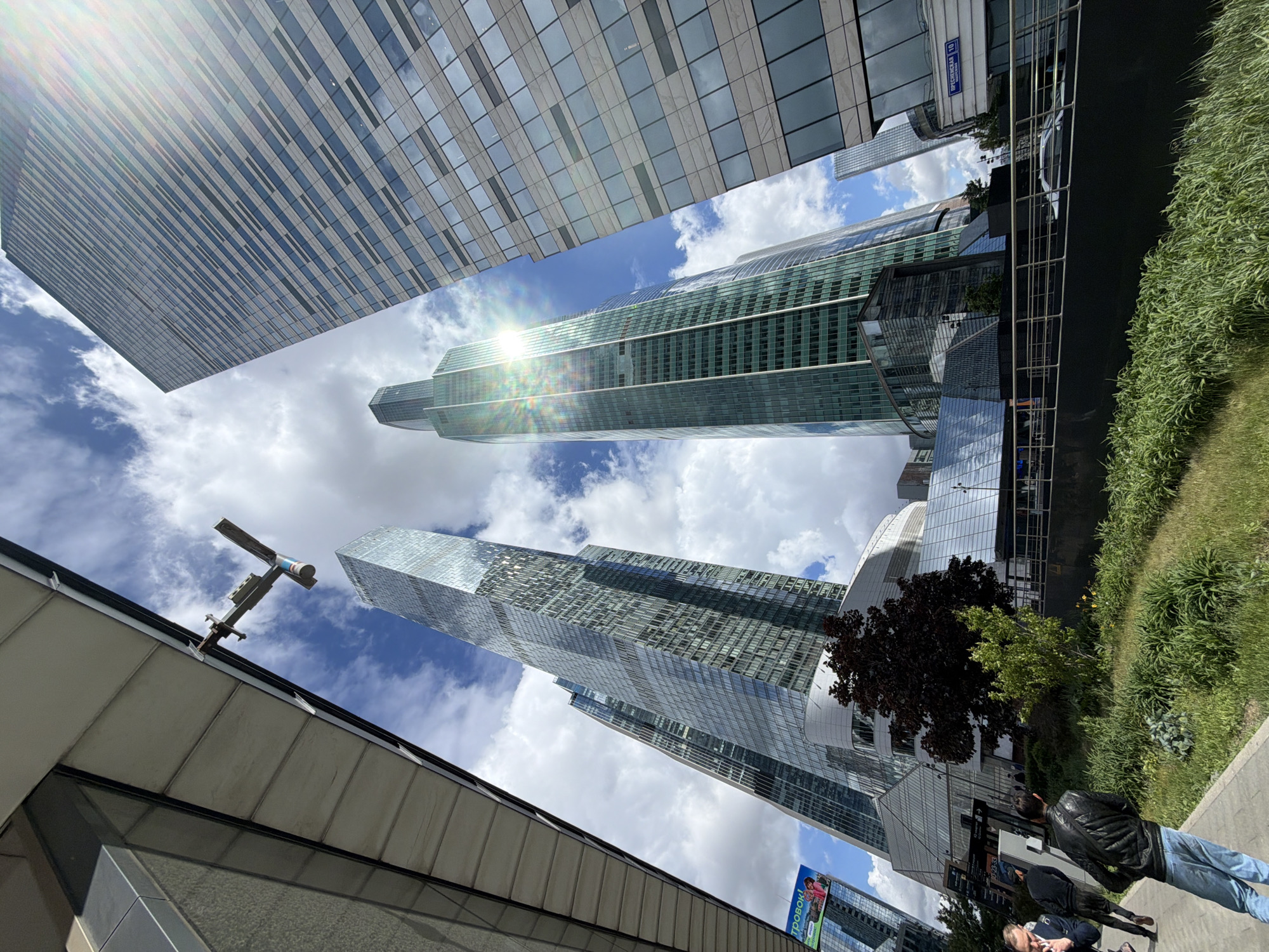 The image shows a view of Moscow International Business Center (Moscow-City) in Moscow, Russia, with several skyscrapers visible against a backdrop of a partly cloudy sky. The architecture is modern, with glass and steel structures. In the foreground, a section of a building or walkway is visible, suggesting the perspective is from street level looking upwards. On the right side of the frame, there's a sidewalk with grass and plants, and two individuals are walking, one in a dark jacket and jeans. A street sign is visible.