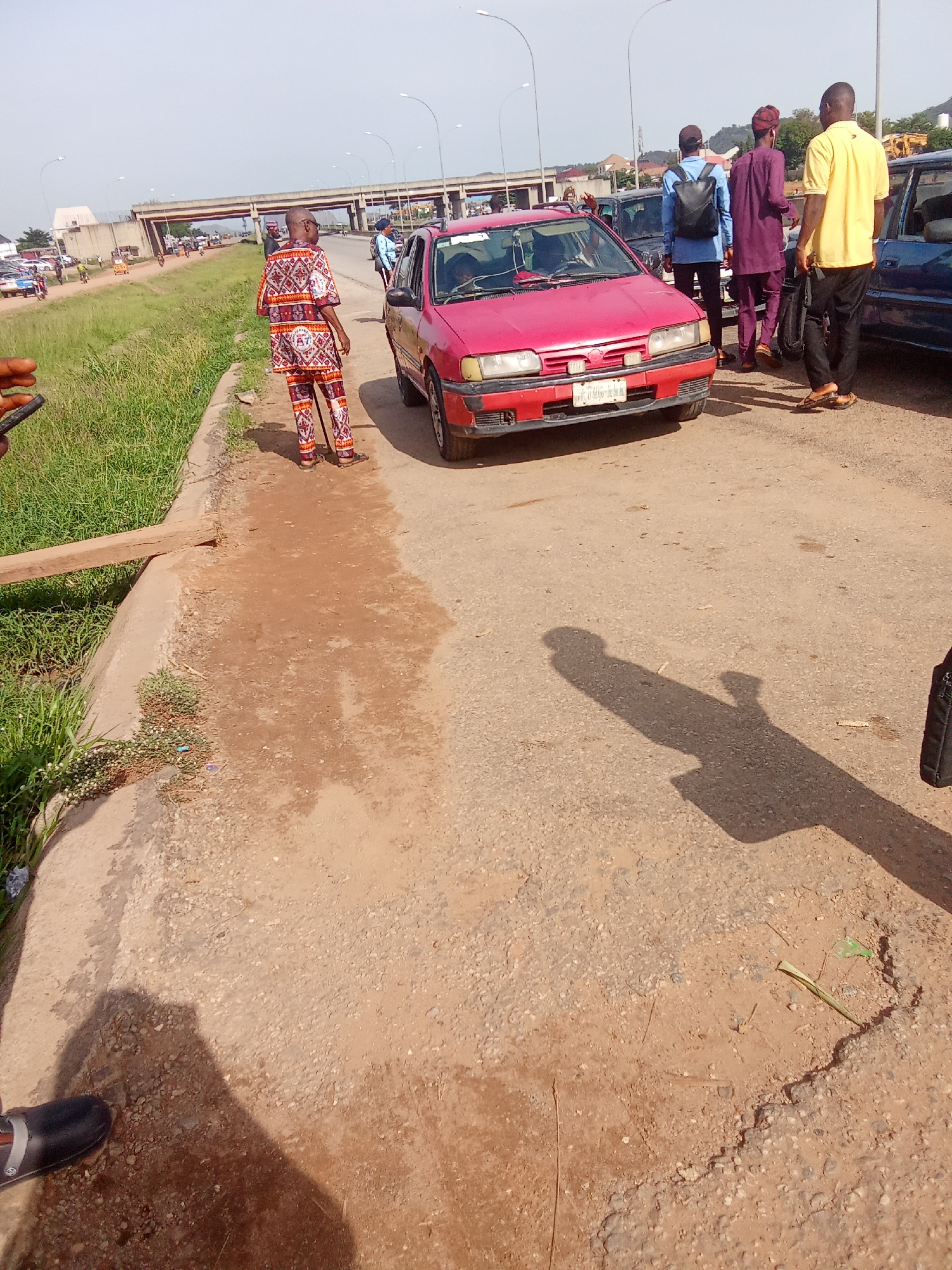 A reddish-pink sedan is parked on a dirt and asphalt road in Kagini, Nigeria. The car is slightly angled towards the viewer, with its front bumper and headlights visible. A man wearing a patterned outfit and sandals stands to the left of the car, facing away from the camera. To the right of the car, three men are standing. The man closest to the car wears a yellow shirt and dark pants. Behind him, a man in a purple outfit and a red headwrap is visible. The third man, furthest back, wears a blue shirt and a black backpack. The background shows a highway overpass, a grassy embankment, and other vehicles. The sky is bright and clear, suggesting it is daytime. The overall scene depicts a roadside gathering or a traffic stop.