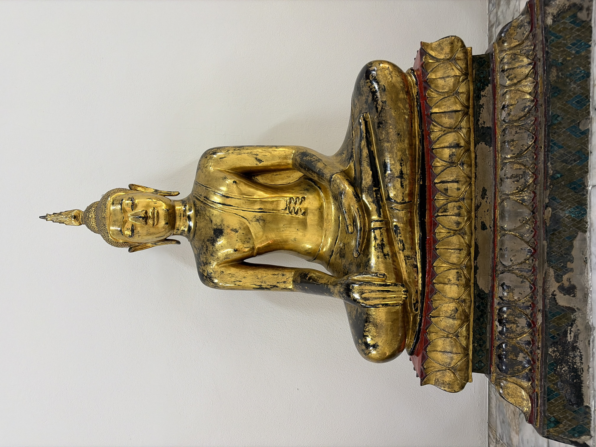 A full-frame, eye-level, medium shot captures a golden Buddha statue seated on an ornate, multi-tiered pedestal against a plain white wall. The statue is predominantly gold with some black detailing, and it appears to be an antique, with some wear and tear visible on its surface. The Buddha is in a meditative pose, with its legs crossed and hands resting in its lap. The pedestal is richly decorated with intricate carvings and patterns, primarily in gold, red, and black, with a mosaic-like pattern on the lowest tier. The floor beneath the pedestal is made of marble tiles. The lighting is even, with no strong shadows, suggesting an indoor setting, possibly a temple or museum. Based on the style of the statue and the typical environment it would be found in, this image is likely taken in Bangkok, Thailand, at a temple such as Wat Phra Chetuphon (Wat Pho), known for its impressive collection of Buddha statues.