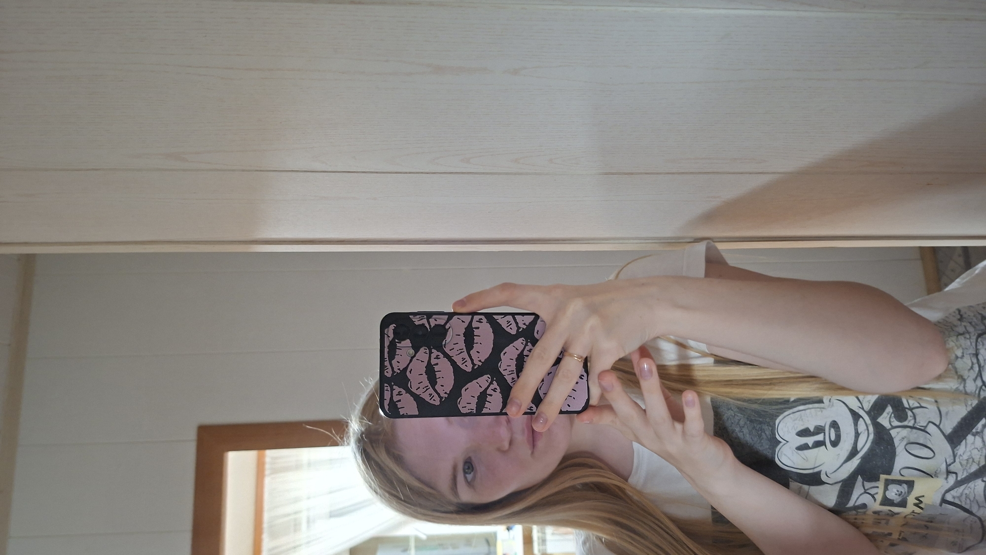 A young woman with long blonde hair takes a selfie in a mirror. She is wearing a white t-shirt with a graphic of Mickey Mouse on it and holding a phone with a pink and black lip pattern case. The background shows a wall with vertical wooden paneling and a window with sheer curtains. The lighting suggests an indoor setting, possibly a home. There is no indication of the specific location within Bolshoy Bukor, Russia.