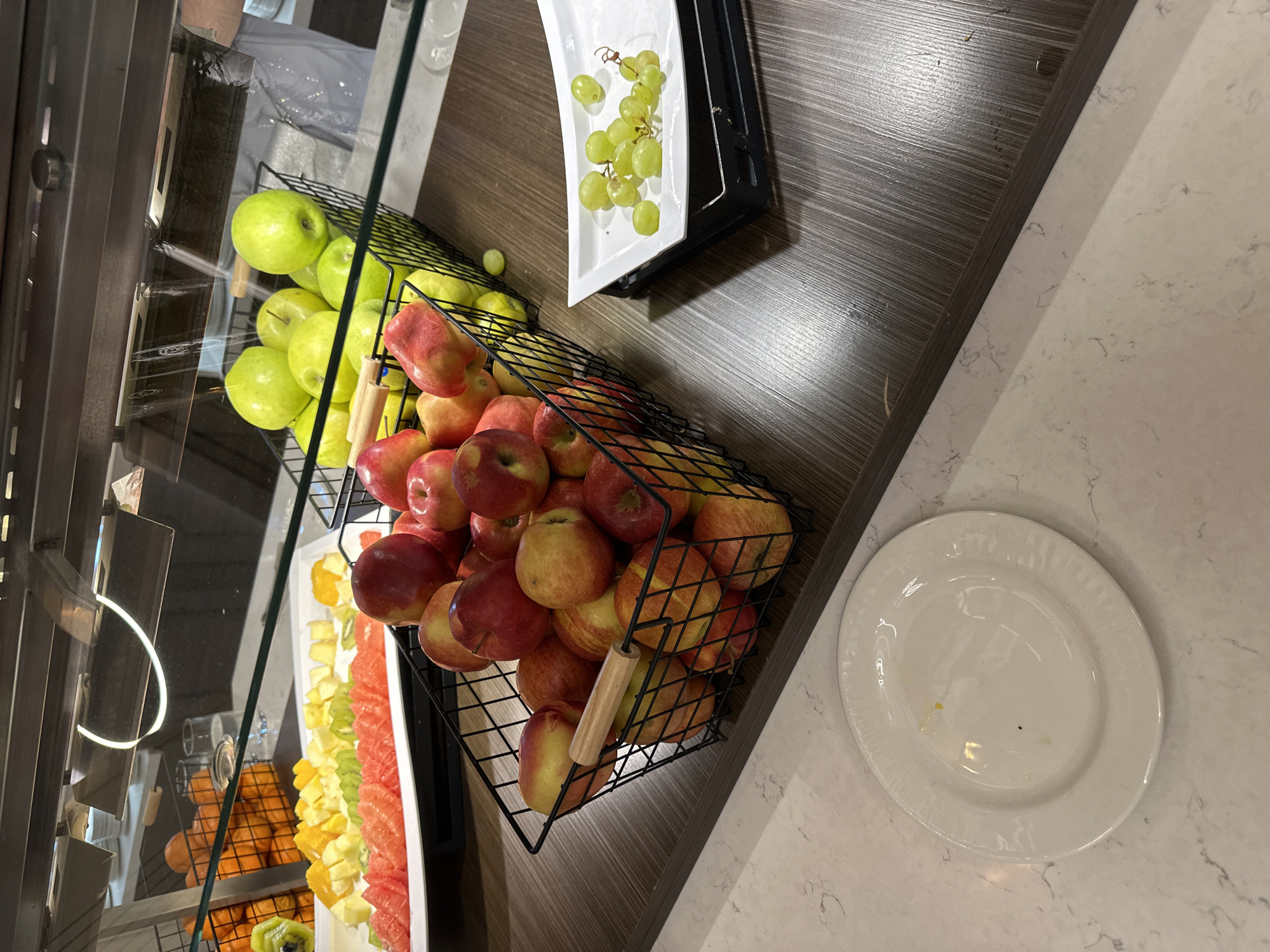 The image shows a fruit display at a buffet. The perspective is a high-angle view, looking down on the display. The display contains different types of fruits. There are green apples and red apples in wire baskets. A long white tray contains sections of cut fruit, including watermelon, kiwi, and oranges. Another white tray holds green grapes. A white plate sits empty next to the basket of red apples. The fruit display is situated on a counter with a wood-grain top and a marble-like countertop. The location is likely a restaurant or hotel. It is not possible to provide the exact location as Kazanlak, Bulgaria.