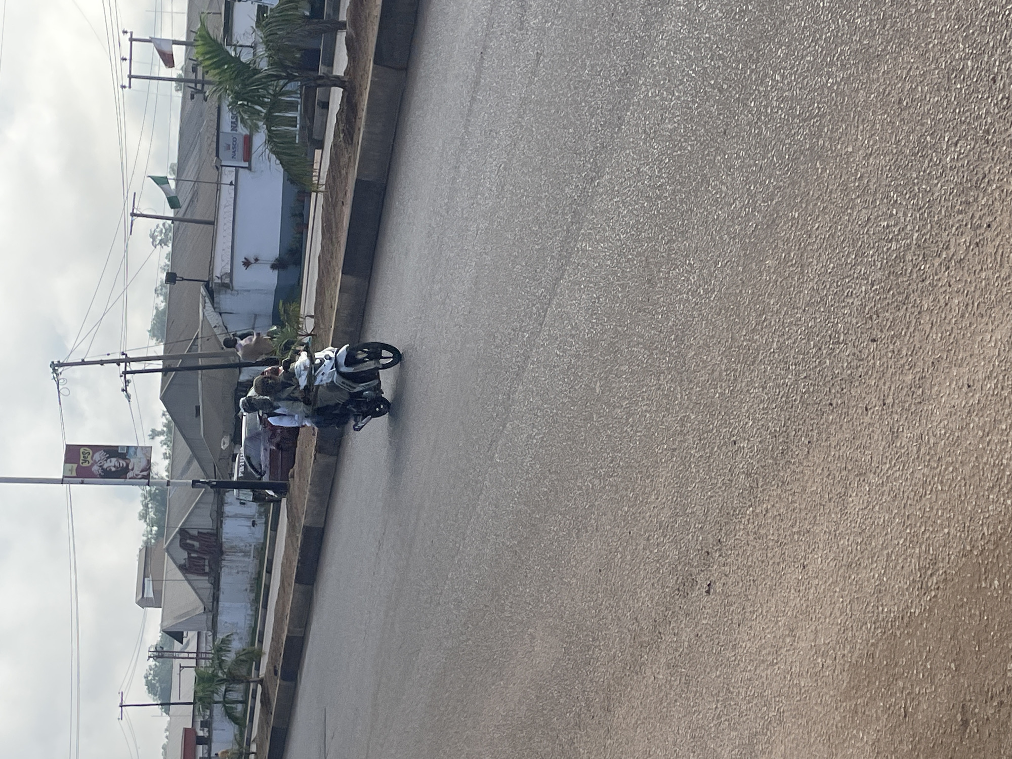 In Jos, Nigeria, a motorcycle carries two individuals down a paved road. The rider, facing left, wears a dark shirt and a head covering. The passenger, also facing left, is attired in a light-colored shirt and a dark head covering. Both appear to be wearing face masks. The motorcycle is white with black accents and is in motion. In the background, buildings with grey roofs are visible, some with white walls. A red car is parked on the side of the road. Several palm trees are planted along the roadside, and a street lamp stands prominently. A banner with an image of a woman and the text "Yes!" hangs from a pole. Another banner features the text "NASCO NASCO" and the company logo, with Nigerian and red flags flown nearby. The sky is overcast. The scene depicts a street in a city, likely during daylight hours.
