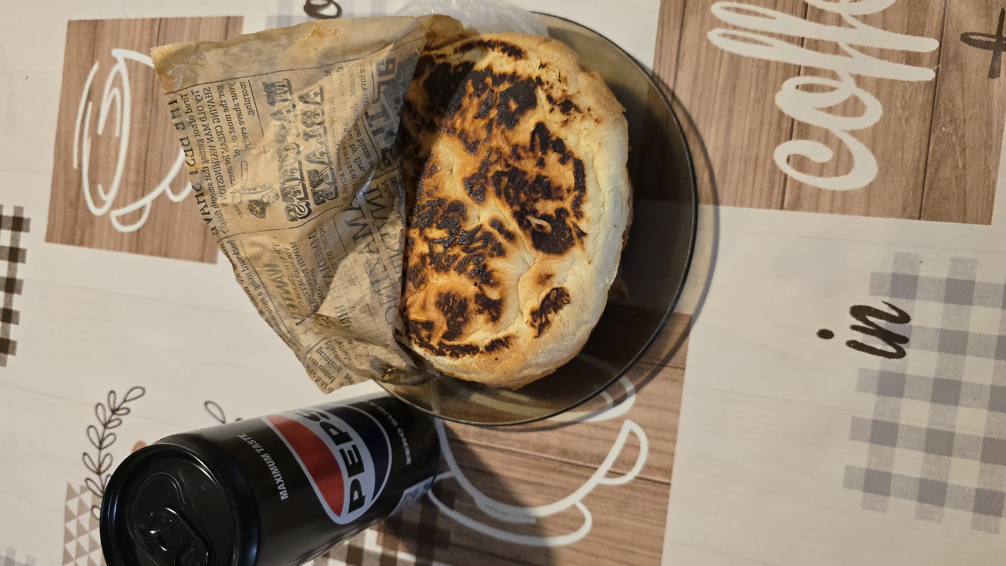 The image shows a close-up, top-down view of a Pepsi can and a piece of bread on a table. The bread, which appears to be a rustic loaf, is partially wrapped in paper that has text printed on it. The bread has a golden-brown crust with some darker, charred areas. The Pepsi can is black with the iconic Pepsi logo. The table is covered with a patterned cloth. In the background, there are subtle decorative elements on the cloth, resembling coffee cups and text. The lighting suggests an indoor setting, likely a home or a cafe, with natural light possibly contributing. There are no people visible in the image. The scene is static, focusing on the food and drink. The specific location within the city of Unknown is not discernible from the image.