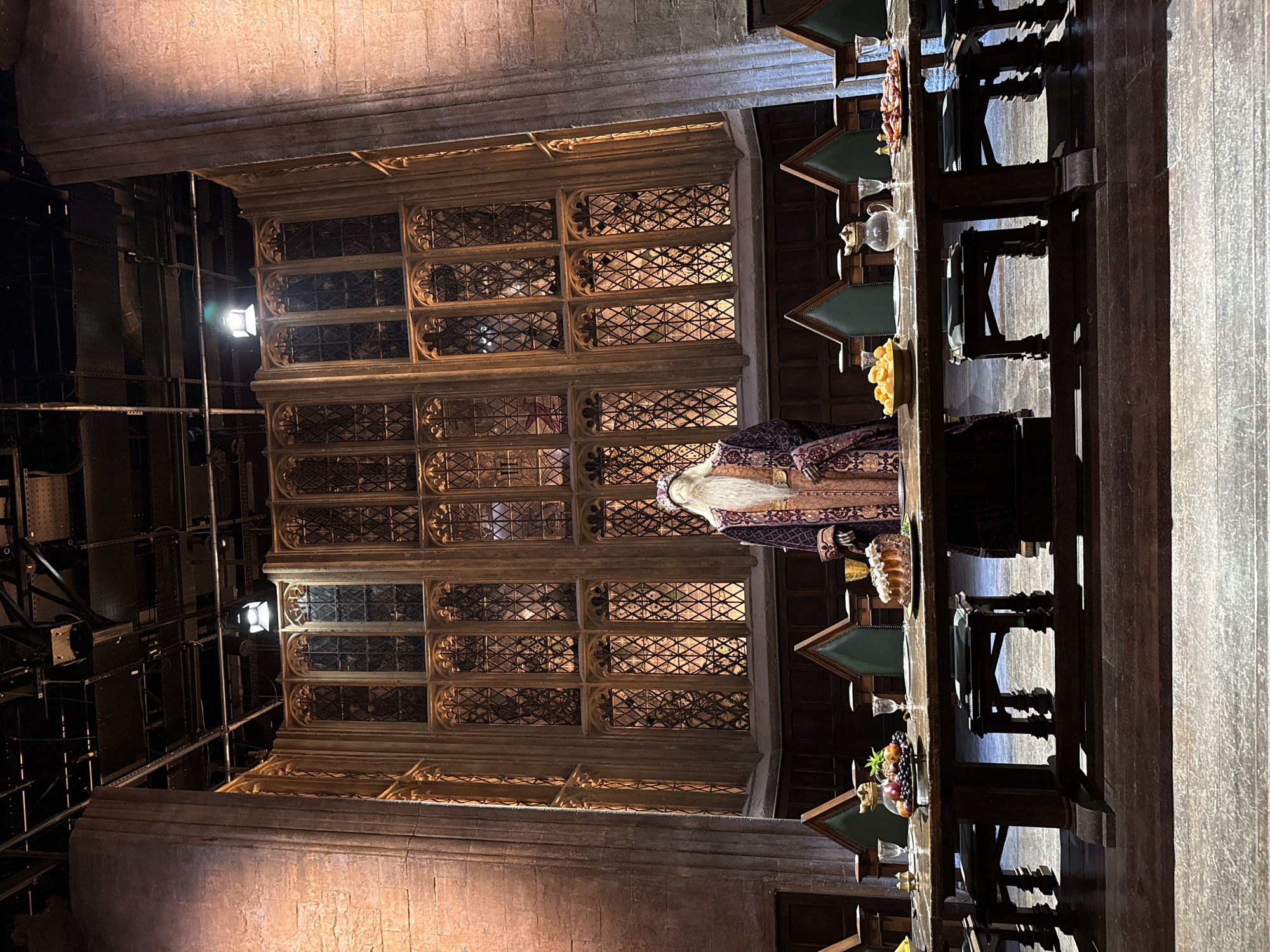 The image depicts the Great Hall set from Warner Bros. Studio Tour London – The Making of Harry Potter in Watford, United Kingdom. A mannequin dressed as Professor Albus Dumbledore, wearing a purple robe with gold embroidery and a long white beard, stands at the head of a long wooden table. The table is laden with food items, including a cake decorated with cream, a bowl of fruit, a platter of what appears to be meats, and a jug of water with glasses. Behind the table, several high-backed green upholstered chairs are arranged. The backdrop features a grand architectural setting with tall stone columns and a large, ornate window with diamond-shaped panes, illuminated from behind. The ceiling above shows exposed theatrical lighting and rigging. The lighting in the scene is warm and atmospheric.