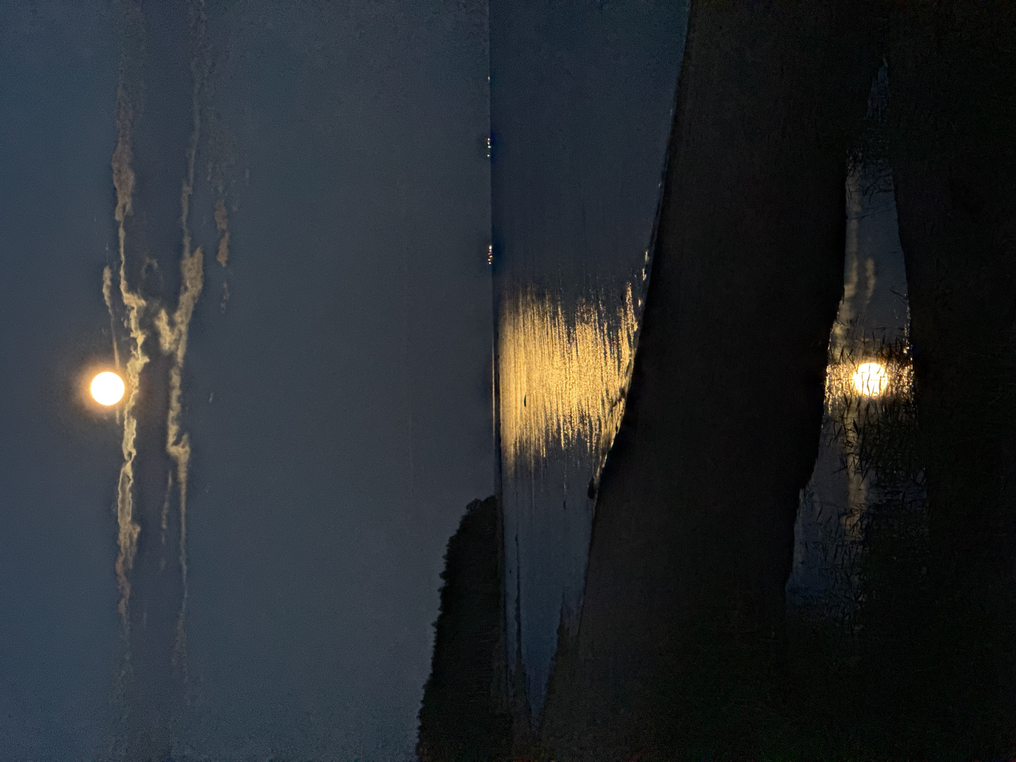 The image depicts a nighttime scene on a beach in Zelenaya Roshcha, Russia. A bright full moon illuminates the sky, partially obscured by scattered clouds. The moonlight casts a shimmering golden reflection on the calm sea, creating a pathway of light. A dark, tree-lined shore is visible to the left, with a sandy beach in the foreground. A small pool of water on the beach reflects the moon and clouds. In the distance, faint lights suggest the presence of ships on the water. The overall atmosphere is serene and tranquil.