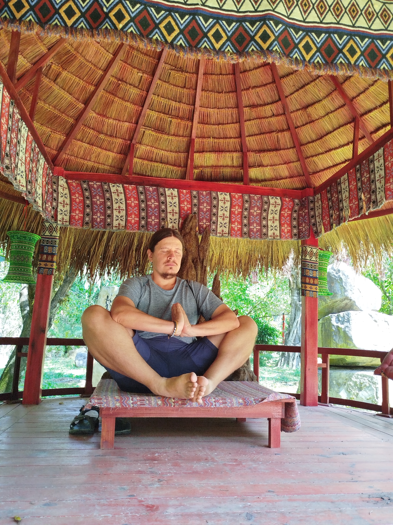 A man is depicted meditating in a tranquil, open-sided gazebo or hut, likely situated in Păng Tiêng 1, Vietnam. He sits cross-legged on a low, patterned wooden platform, with his eyes closed and hands pressed together in a prayer position (Anjali Mudra). He wears a grey t-shirt, dark shorts, and a bracelet on his right wrist. His black sandals are placed on the weathered wooden floor next to the platform.
The gazebo features a rustic thatched roof, supported by red-painted wooden beams and posts. Colorful, intricately patterned fabrics or tapestries adorn the structure, draped along the roofline and wrapped around some of the support pillars. These decorative elements add a vibrant and traditional aesthetic to the setting.
Beyond the gazebo, lush green foliage and large, light-colored boulders are visible, indicating a natural and possibly tropical outdoor environment. The bright, soft lighting suggests it is daytime, perhaps late morning or early afternoon, with clear weather. The man's posture and closed eyes convey a sense of calm, focus, and serenity, consistent with meditation. There is no visible text in the image.