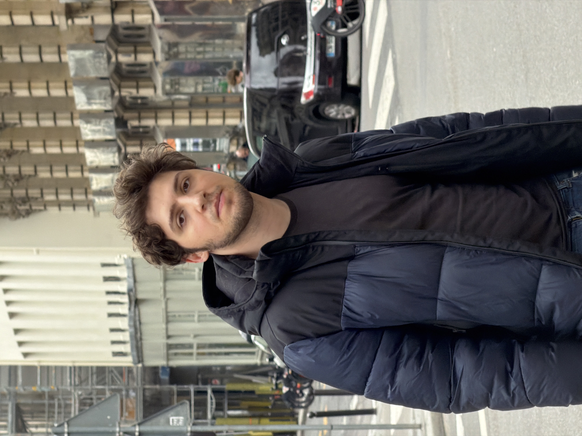 The image shows a man with brown, curly hair and a beard, wearing a dark-colored puffer jacket and a black t-shirt. He is standing in an urban environment, likely in Paris, France. The background includes buildings with architectural details and metal structures. There is a black car and a bicycle parked in the background, suggesting the setting is a street. The man is looking directly at the camera. There is sunlight present. The image orientation is vertical. The environment includes pedestrian crosswalk markings on the street.