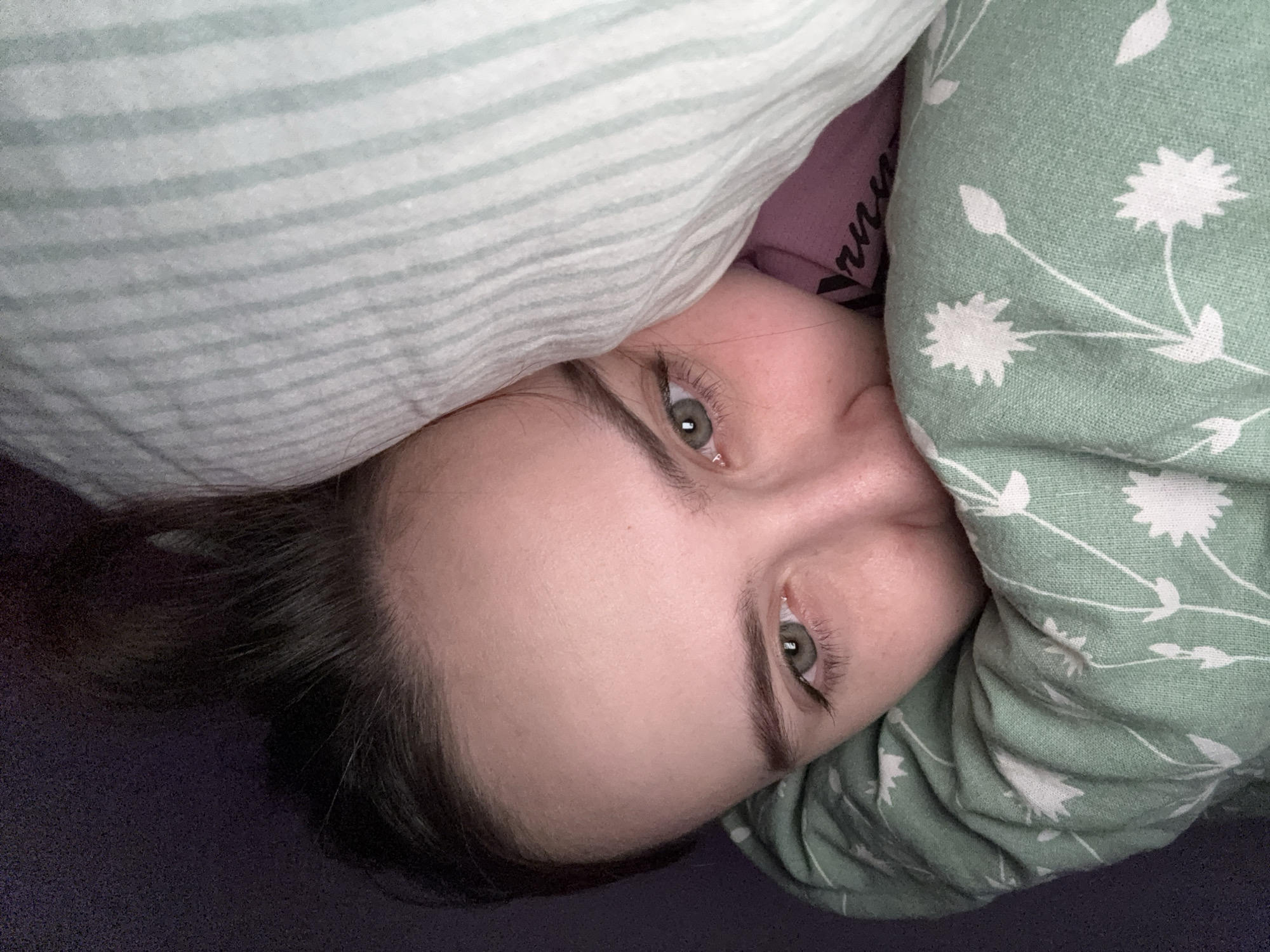 A close-up shot depicts a person lying in bed, partially covered by bedding. The individual has fair skin, green eyes, and dark hair. One side of the face is partially obscured by a green blanket patterned with white floral designs. The other side of the face is near a striped white and light green blanket. The person appears to be in a relaxed or resting state. The background is a dark, solid color, likely the bed itself. It is impossible to verify that this photo was taken in Прикамский, Russia.