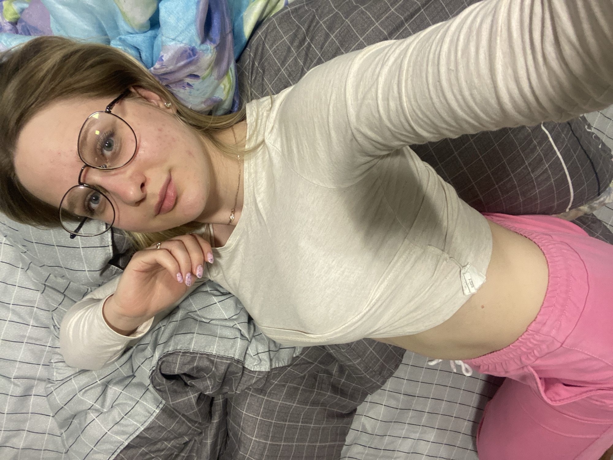 A young woman with blonde hair and round glasses is lying on a bed with gray and blue checkered bedding. She is wearing a long-sleeved white shirt and pink sweatpants. Her right arm is extended towards the camera. Her left hand is resting on the bed next to her head. No location information is included in the image.