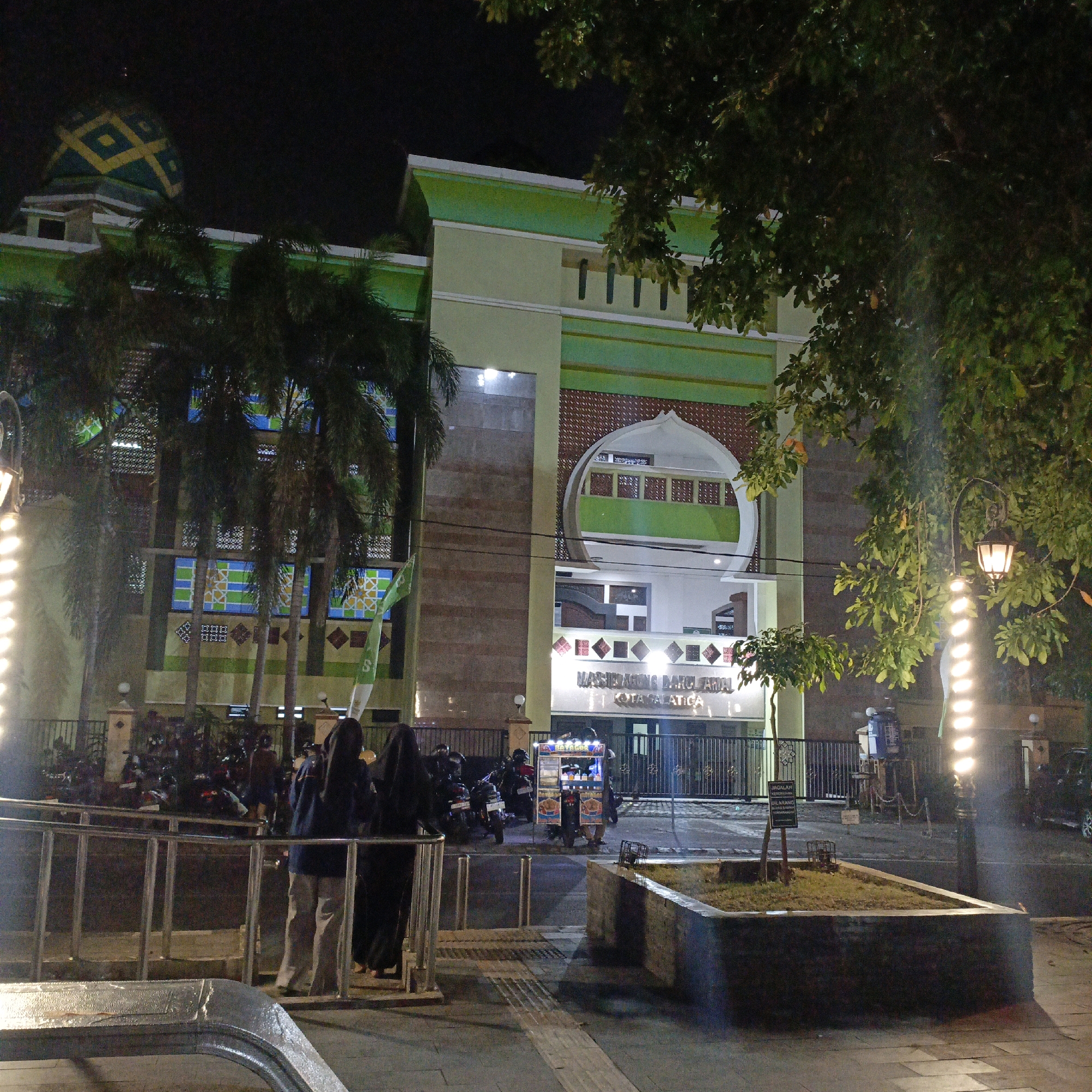 A nighttime scene unfolds in front of a mosque in Salatiga, Indonesia. The mosque, with its distinctive green dome adorned with yellow geometric patterns, is illuminated, casting a warm glow. The building's facade features a light green color with white trim, and decorative patterned panels in shades of blue, green, and brown. Above the main entrance, a white arched structure is visible, housing a green balcony with intricate latticework.
In front of the mosque, several palm trees stand tall, their fronds creating a lush silhouette against the night sky. A street lamp with a decorative black lantern and glowing bulbs adds to the ambiance. A small planter box with sparse vegetation is situated in the foreground.
Several people are visible, including two women in dark clothing standing near a metal railing, and a group of people and motorcycles parked further back. A food cart with a colorful display is also present. The ground is paved with patterned tiles, and a sidewalk is visible.
A sign with Indonesian text is posted on a small stake in front of the planter box, although the text is difficult to read clearly. Another sign above the mosque entrance reads "MASJID AGUNG DARUL AMAL KOTA SALATIGA," identifying it as the Darul Amal Grand Mosque in Salatiga. The overall atmosphere is peaceful and serene, suggesting a typical evening scene in a community gathering place.