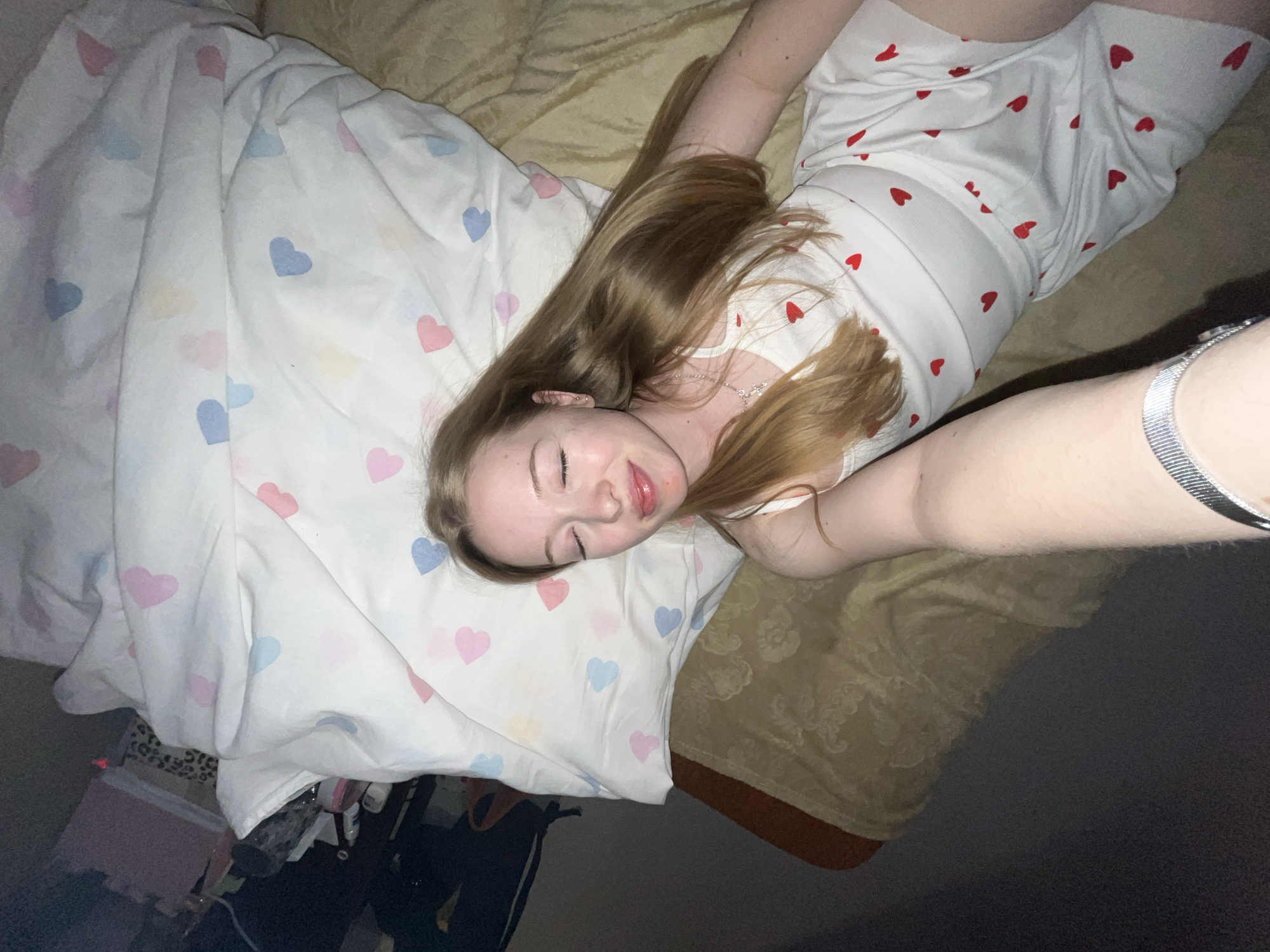 An overhead, slightly angled shot shows a young individual, likely female, resting or asleep on a bed in an indoor setting in Chaikovsky, Russia. The person, with long, light brown hair, is lying with closed eyes and a relaxed expression, head on a white pillow adorned with pastel pink, blue, yellow, and purple heart patterns.
The individual is wearing a white top or dress featuring small red heart patterns. A delicate silver-colored chain necklace with a small pendant is visible around their neck. On their right wrist, a wide, silver-colored bracelet or watch is worn.
The bed is covered with a textured gold-colored bedspread or blanket. The surroundings beyond the bed are dimly lit, showing dark flooring or carpet. In the lower left corner, partially obscured objects on the floor include a pink box or container, a leopard print item, and possibly a bottle. The overall impression is one of peaceful rest. There is no visible text in the image.