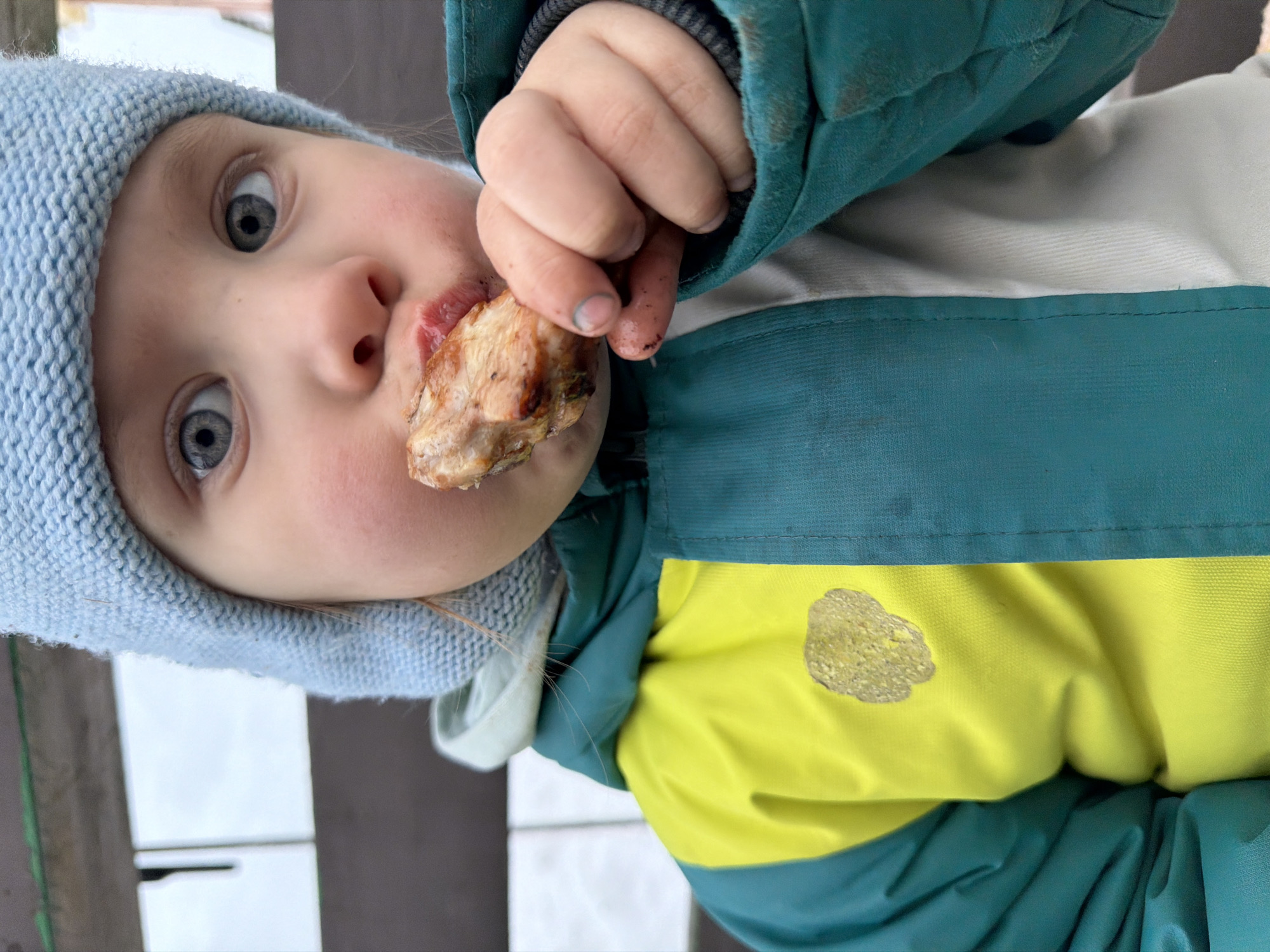 The image presents a close-up, chest-up view of a young child, approximately two to five years old, engaged in eating outdoors in the city of Chaikovsky, Russia. The child possesses fair skin, light blue-gray eyes, and wears a light blue knit beanie. The upper body is clad in a winter jacket featuring a teal lower and sleeve section and a bright yellow upper chest section. A light-colored, possibly white or light grey, layer is visible beneath the jacket. The child's mouth is partially open, actively biting into a piece of cooked meat, likely a bone-in fragment such as a chicken wing. The child holds the meat with their left hand; visible fingers exhibit dirt and a blue stain on the tip of the index finger. A soiled, textured patch is evident on the yellow part of the jacket's chest area. The background is out of focus, revealing a dark, indeterminate structure to the right and a lighter, rectangular surface contrasting with a bright, overcast sky to the upper left.