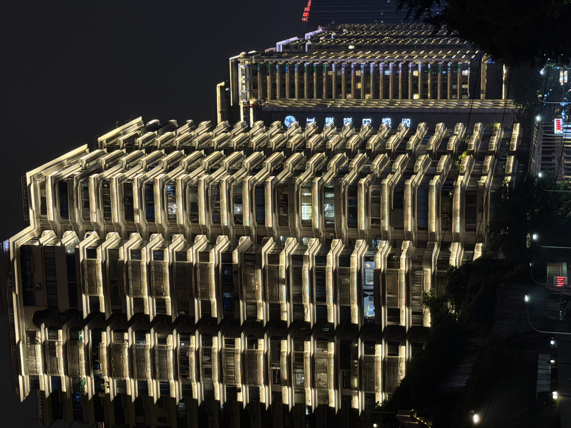 The image shows a nighttime view of modern apartment buildings in Yuzhong District, China. The buildings are tall and feature a distinctive architectural style with numerous balconies and tiered structures. Warm yellow lights illuminate the exterior of the buildings, outlining the balconies and the edges of the floors, creating a striking visual effect against the dark night sky. The buildings are densely packed, suggesting an urban environment. In the background, another taller building is visible with some red neon signage, and to the right, some trees are present. The lower part of the image shows streetlights and some indistinct structures, indicating a street level view. The scene conveys a sense of urban density and illuminated modernity.