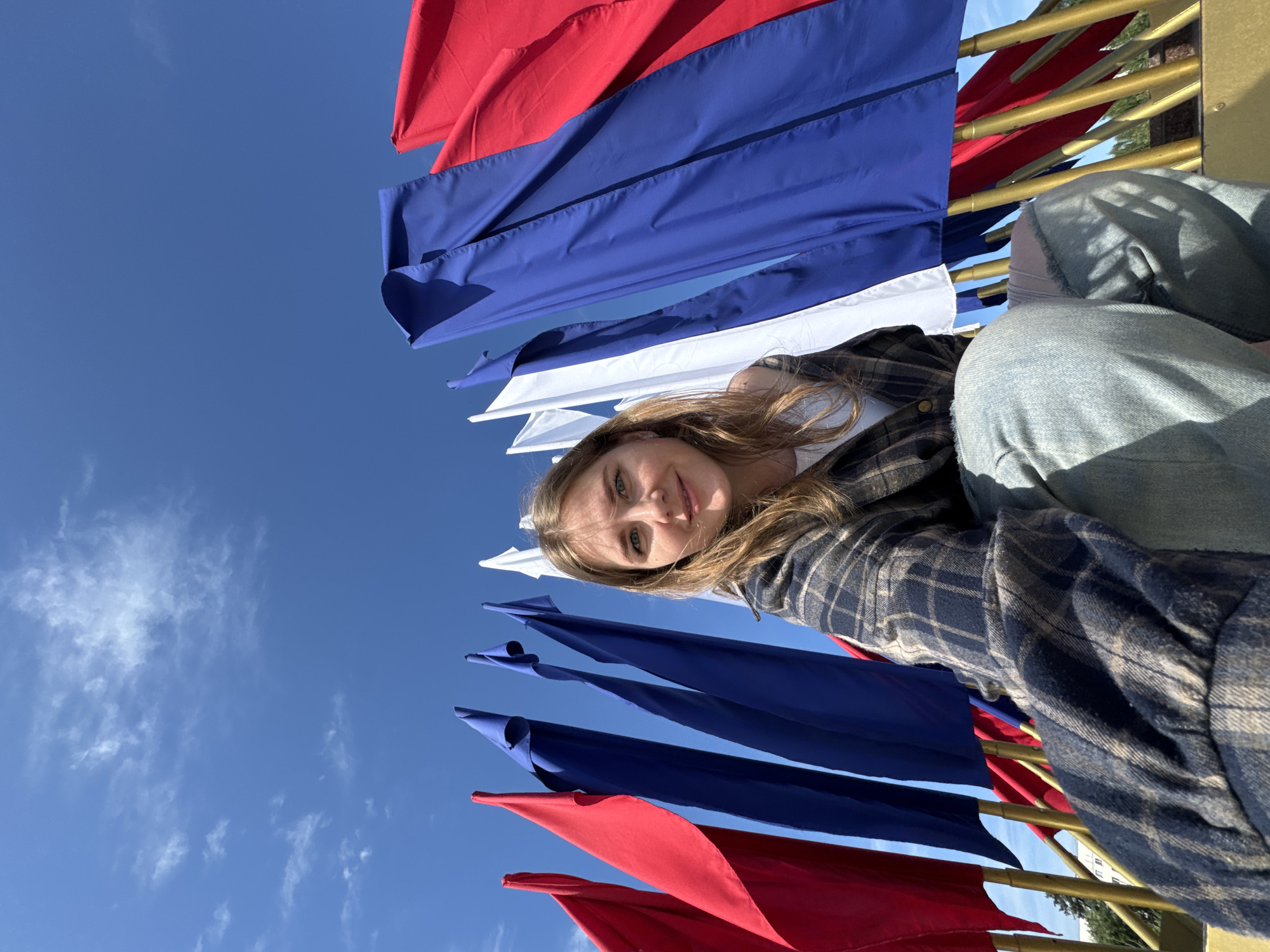 A young woman is captured in a close-up portrait, looking directly at the camera with a gentle smile. She has long, light brown hair cascading around her face and is wearing a dark-colored plaid shirt and blue jeans, partially visible on the right. The image is taken from a low angle, looking upwards towards a bright, clear blue sky with sparse, wispy white clouds.
Behind and around the woman, numerous banners or flags in red, blue, and white are prominently displayed, suggesting the colors of the Russian flag (white, blue, red). These flags hang vertically and appear to be attached to golden or brass-colored poles or railings, which are also visible. The arrangement of the flags suggests they are part of a decorative display or celebration. The bright sunlight and clear sky indicate it is daytime, likely a sunny day. The overall impression is one of a casual, outdoor setting, possibly a public space in Perm, Russia, given the provided context. There is no visible text in the image.