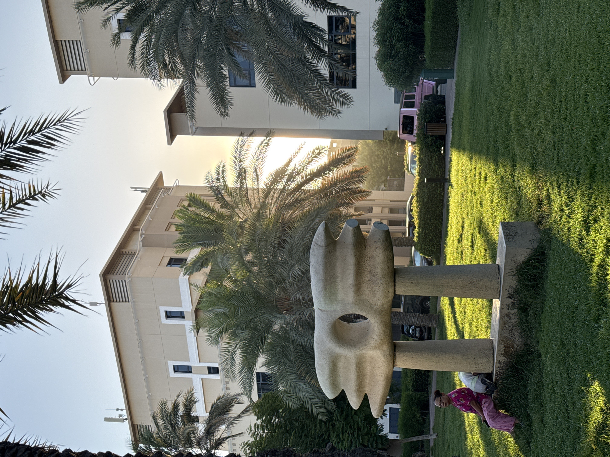 In Dubai, United Arab Emirates, an outdoor scene unfolds in the warm glow of sunset. In the foreground, a sculpture made of light-colored stone, supported by two cylindrical pillars, stands on a concrete base in a grassy park. To the left of the sculpture, a woman wearing a pink top and pink pants sits on the concrete base, facing left. Next to her, a white object, possibly a bag or a pillow, rests on the ground. The park features lush green grass, and palm trees with long fronds are scattered throughout the area. In the background, two modern beige buildings with multiple floors and windows rise into the sky. Between the buildings, the setting sun casts a warm, golden light. A pink SUV is parked behind a hedge in front of one of the buildings. The overall impression is one of a tranquil residential area in Dubai bathed in the soft light of the evening.