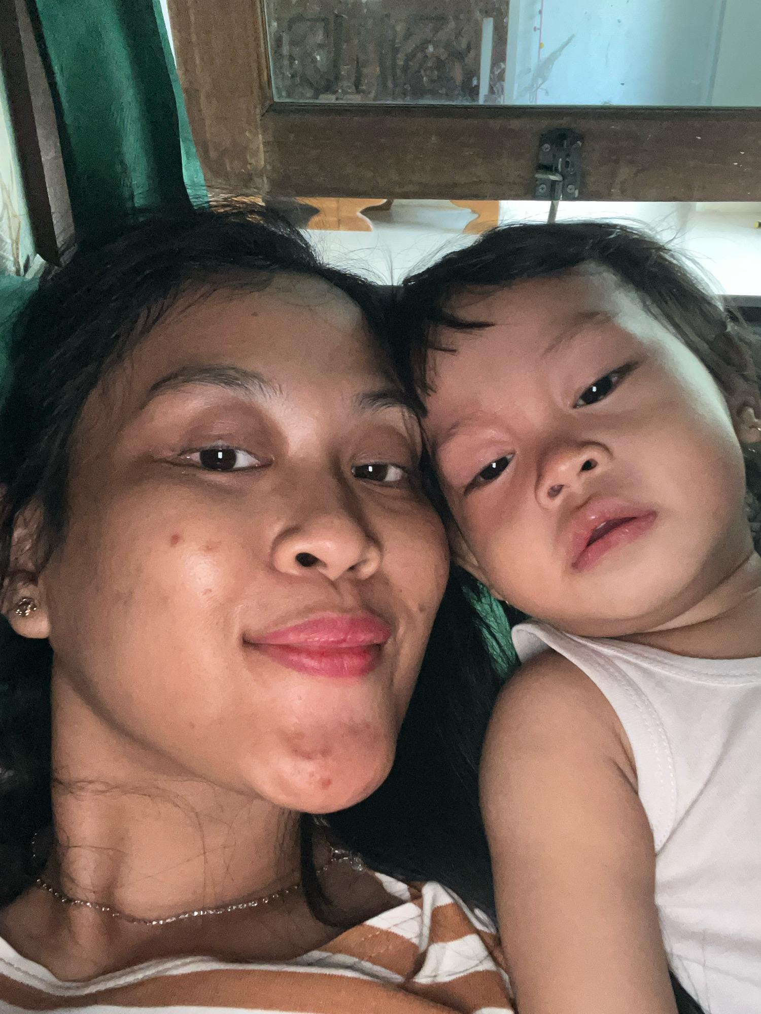 This is a close-up, selfie-style photograph of a young Asian woman and a toddler, positioned side-by-side and looking towards the camera. The woman is on the left, with her dark hair framing her face. She has a light complexion and is wearing a necklace. Her lips are slightly parted, and she is smiling. The toddler, on the right, has a round face and dark hair. They are wearing a white tank top. Their expression is neutral, with their eyes looking towards the camera.
The background is blurred, but suggests an indoor setting. To the left, there is a dark green curtain. To the right, a wooden frame is visible, with a metal latch attached to it. The lighting appears to be artificial, casting a warm glow on their faces. The image quality is slightly grainy. The presence of a window or door frame in the background might suggest a residential interior, possibly a home in Kecamatan Pati, Indonesia.
