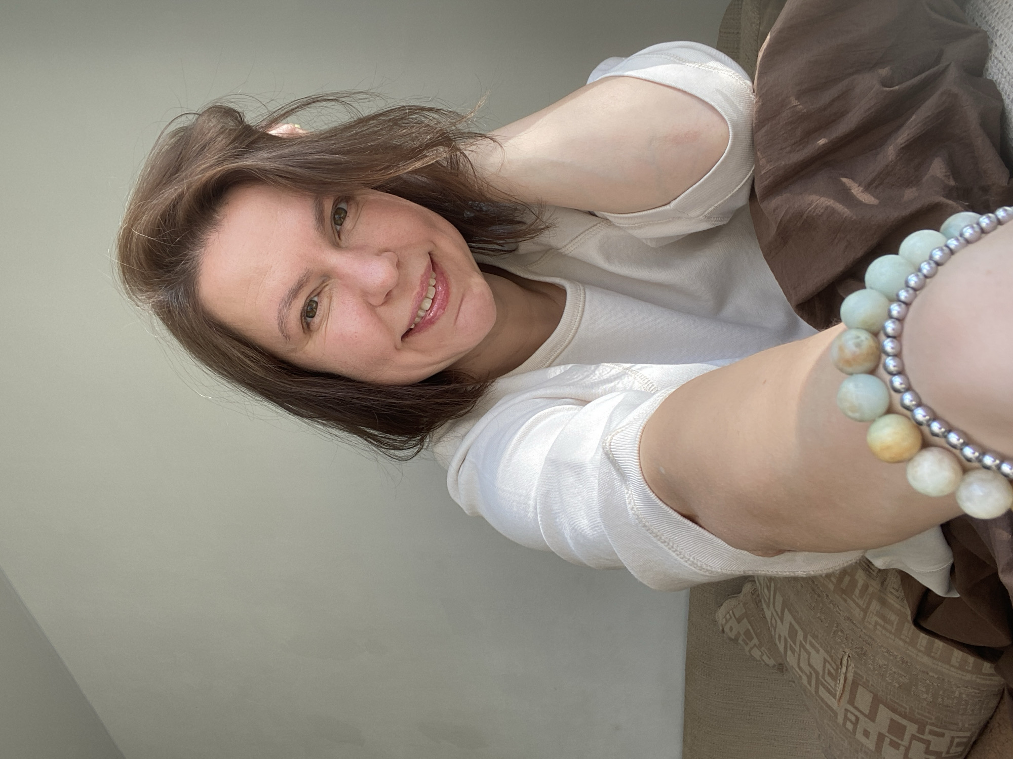 A woman with short brown hair and a light complexion smiles into the camera. She wears a white long-sleeved shirt and a beaded bracelet. She appears to be indoors, near a window providing natural light. A brown garment or fabric is visible behind her arm. A patterned cushion is also partially visible. There is no information in the image to confirm the location in Chaikovsky, Russia.