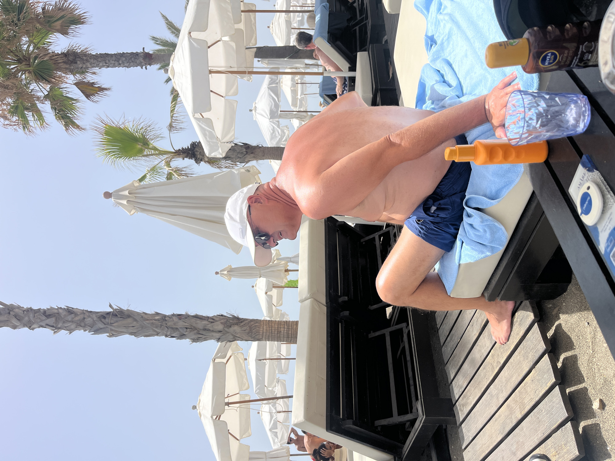 A medium shot captures a man in his 50s, wearing a white baseball cap, sunglasses, and blue swim trunks, sitting on a lounger at a beach resort in Marbella, Spain. He is partially reclined, with his back to the camera, facing towards the right. His skin is visibly sun-tanned and slightly reddened.
In front of him, on a dark wooden table, are several items: an orange sunscreen bottle with a ribbed texture, a clear blue patterned glass, and a dark brown bottle of Nivea Sun tanning oil. A light blue towel is draped over the lounger.
The background reveals a sunny beach scene with numerous white beach umbrellas and palm trees. Other beachgoers are visible in the distance, also relaxing on loungers. The ground is a mixture of sand and wooden decking. The sky is clear and bright blue.