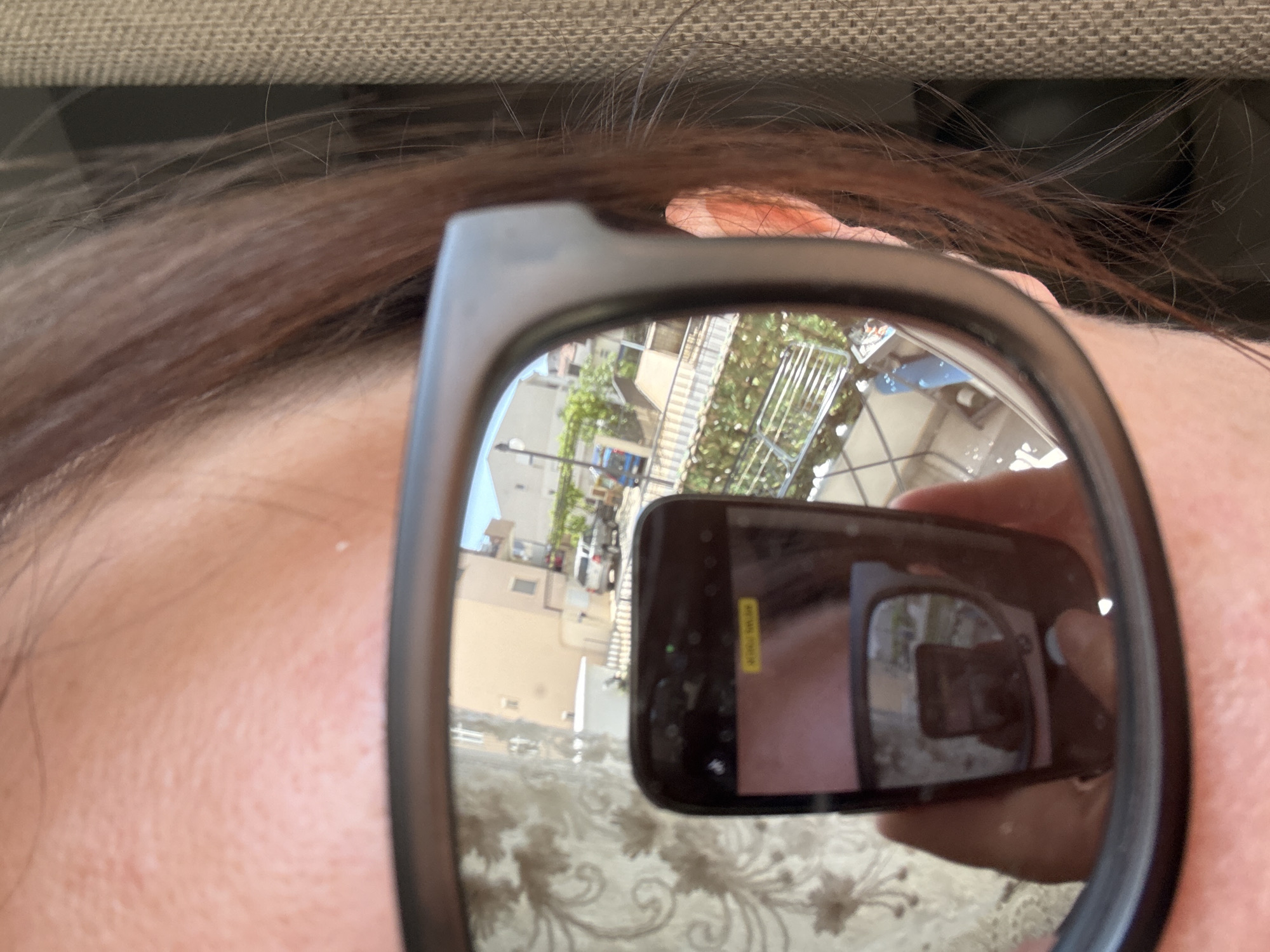 The image shows a person wearing sunglasses, with the reflection in the lenses revealing a cityscape. A hand holding a cell phone is visible in the sunglasses' reflection, indicating the image is a photograph taken with the cell phone. The cityscape in the reflection appears to include residential buildings and greenery, suggestive of an urban or suburban environment. Given the prompt's specification, the location is likely Bar, Montenegro, but this is only deduced information based on the prompt. Specific buildings or landmarks visible in the reflection are not identifiable. The person is close to the camera, partially out of focus, and appears to be indoors. The background behind the person is out of focus and shows a neutral-colored, textured surface possibly belonging to furniture.