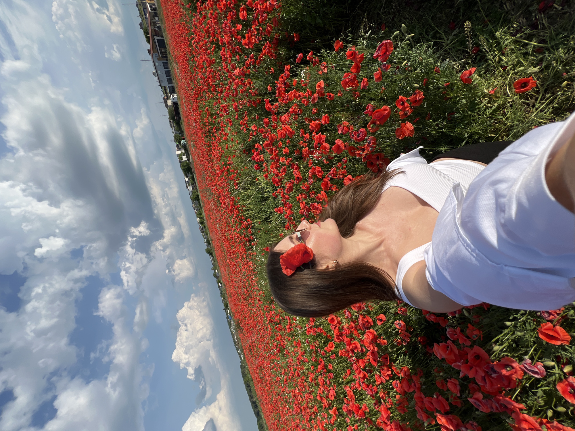 A young woman with brown hair, adorned with a red poppy flower above her right ear, stands in a vast field of red poppies under a partly cloudy sky. She is wearing a white crop top and sunglasses, and holds a white garment draped over her arm. In the background, a gentle slope of buildings and trees can be seen. The scene is reminiscent of the fields that surround the city of Ternopil, Ukraine, known for their stunning natural beauty. The bright red of the poppies contrasts with the green foliage and the soft blue of the sky, creating a vibrant and picturesque landscape. The woman's gaze is directed to her right, her profile captured against the backdrop of the poppy field.