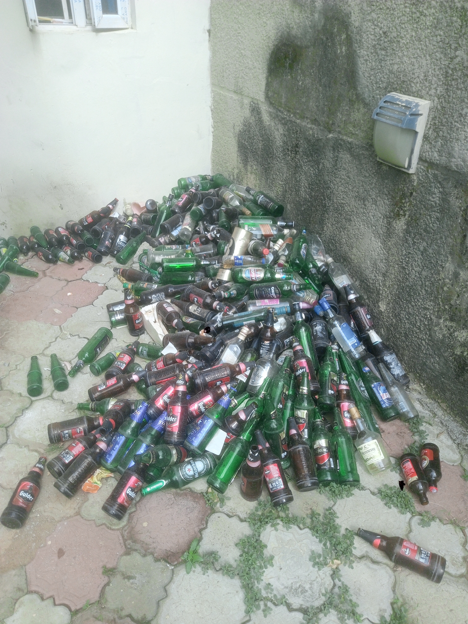 The image depicts a large pile of empty alcoholic beverage containers discarded against a wall. The bottles are primarily glass and include beer bottles of brands such as Gulder, Heineken, and Star, as well as liquor bottles. Some aluminum cans are also visible. The location appears to be outdoors, on a tiled surface that is somewhat overgrown with weeds. The wall is a mix of painted white sections and unpainted textured concrete. There's an outdoor light fixture affixed to the concrete wall. Based on the presence of Gulder beer, a product popular in Nigeria, and the geotag information, it's possible the location is in the city of Kubwa, Nigeria.