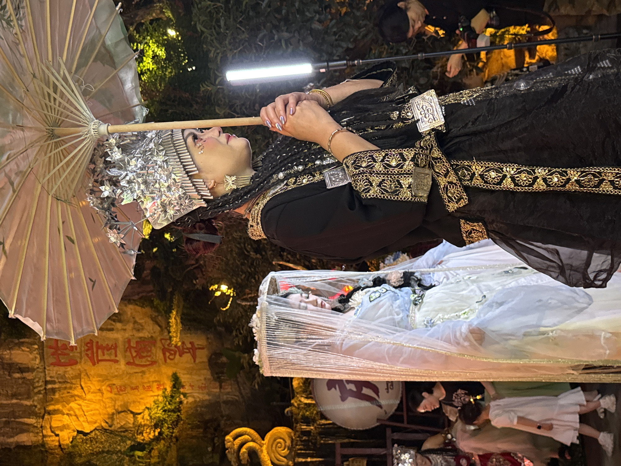 In Yuzhong District, China, at night, a woman in elaborate traditional attire stands holding a parasol. She wears a tall, ornate silver headdress adorned with floral motifs and butterfly earrings. Her black garment is heavily embroidered with gold trim. Next to her, another woman, also in traditional attire, is partially obscured by a sheer veil. She wears a light blue and white layered dress, also adorned with delicate embellishments. In the background, a stone wall is illuminated with warm yellow light, displaying red Chinese characters. To the right, a young girl in a white dress and sneakers walks away from the viewer, while other individuals are present in the dimly lit environment, including a person holding a camera. The scene suggests a cultural performance or celebration.