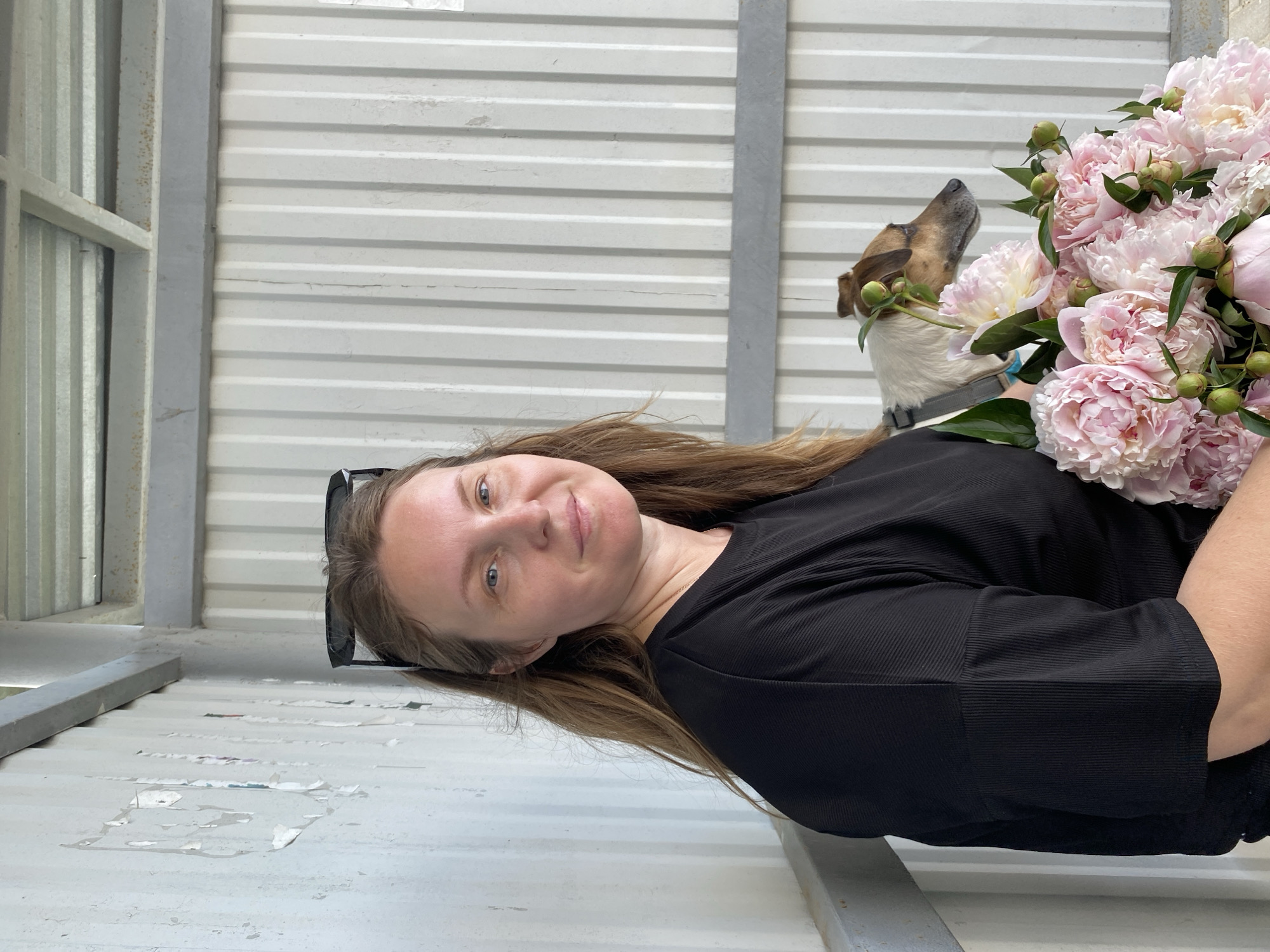 An outdoor shot, likely a covered area given the corrugated metal wall and ceiling, features a woman and a dog. The woman, with long brown hair and blue eyes, wears a black ribbed shirt and has sunglasses on her head. She is holding a bouquet of pink peonies and a small white and brown dog with floppy ears. The dog is wearing a grey harness. The background is a neutral-toned corrugated metal wall and a ceiling structure. The lighting suggests it is daytime.