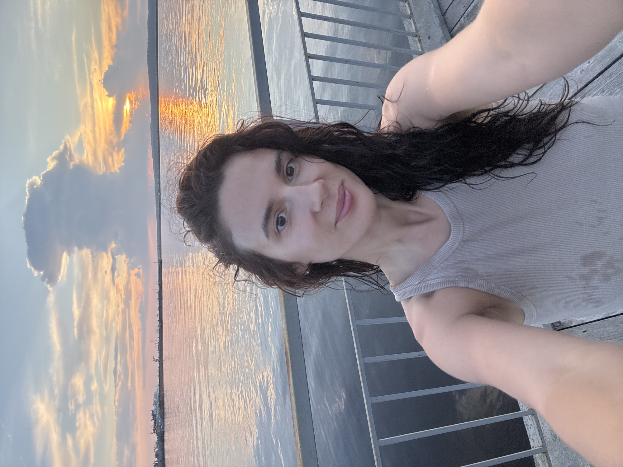 This is a selfie of a woman taken on a pier at sunset. She has long, dark, wavy hair and is wearing a light-colored tank top. The water is calm, reflecting the colors of the sunset, and a distant shoreline with buildings and cranes is visible. The sky is filled with dramatic clouds tinged with orange and pink. The pier is made of wood planks and has a metal railing. The woman is looking directly at the camera with a slight smile. The location is the Kama River embankment in Chaikovsky, Russia, a city known for its industrial port and hydroelectric power station.