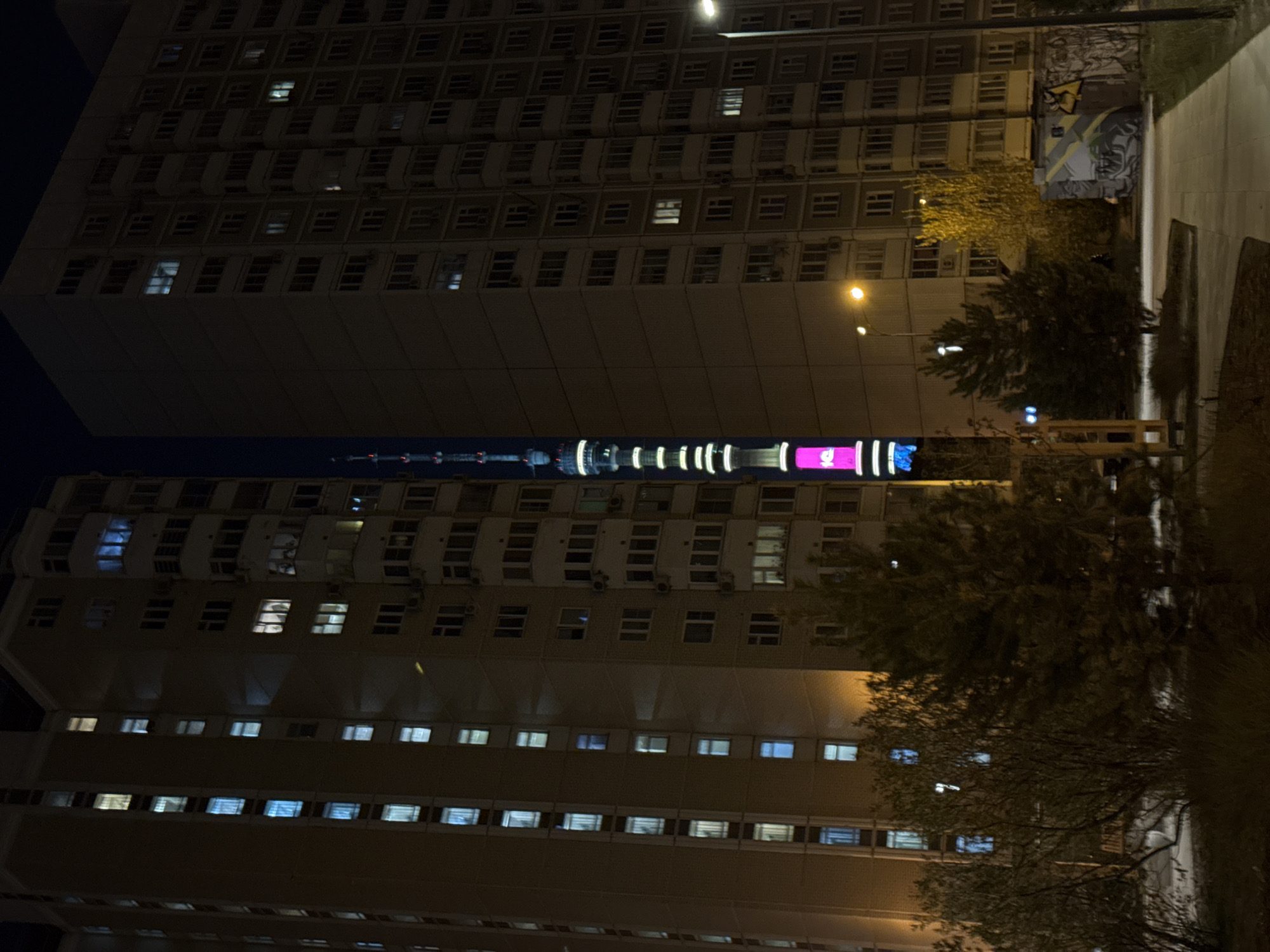At night, a tall residential building stands in Khimki, Russia. The building, composed of multiple stories with numerous windows, partially obscures the Moscow TV tower in the background. Streetlights illuminate the trees and the walkway along the side of the building. There is also a graffiti wall near the side of the building.