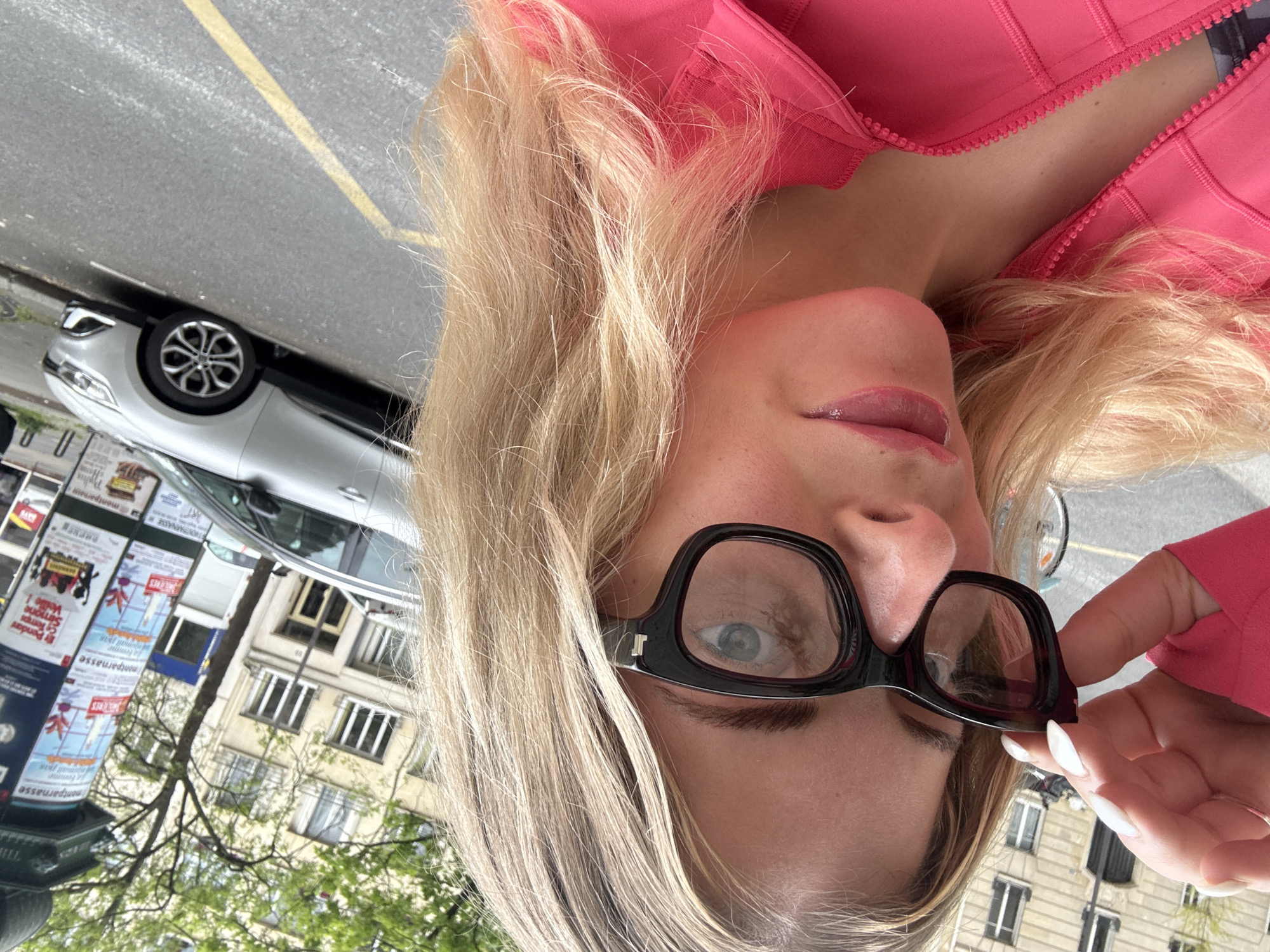 A woman with blonde hair, wearing black-framed glasses and a pink top, is holding the glasses away from her face, presumably taking a selfie. A light-colored vehicle is parked on the street, and in the background are buildings and street signs, indicating a location in Paris, France.