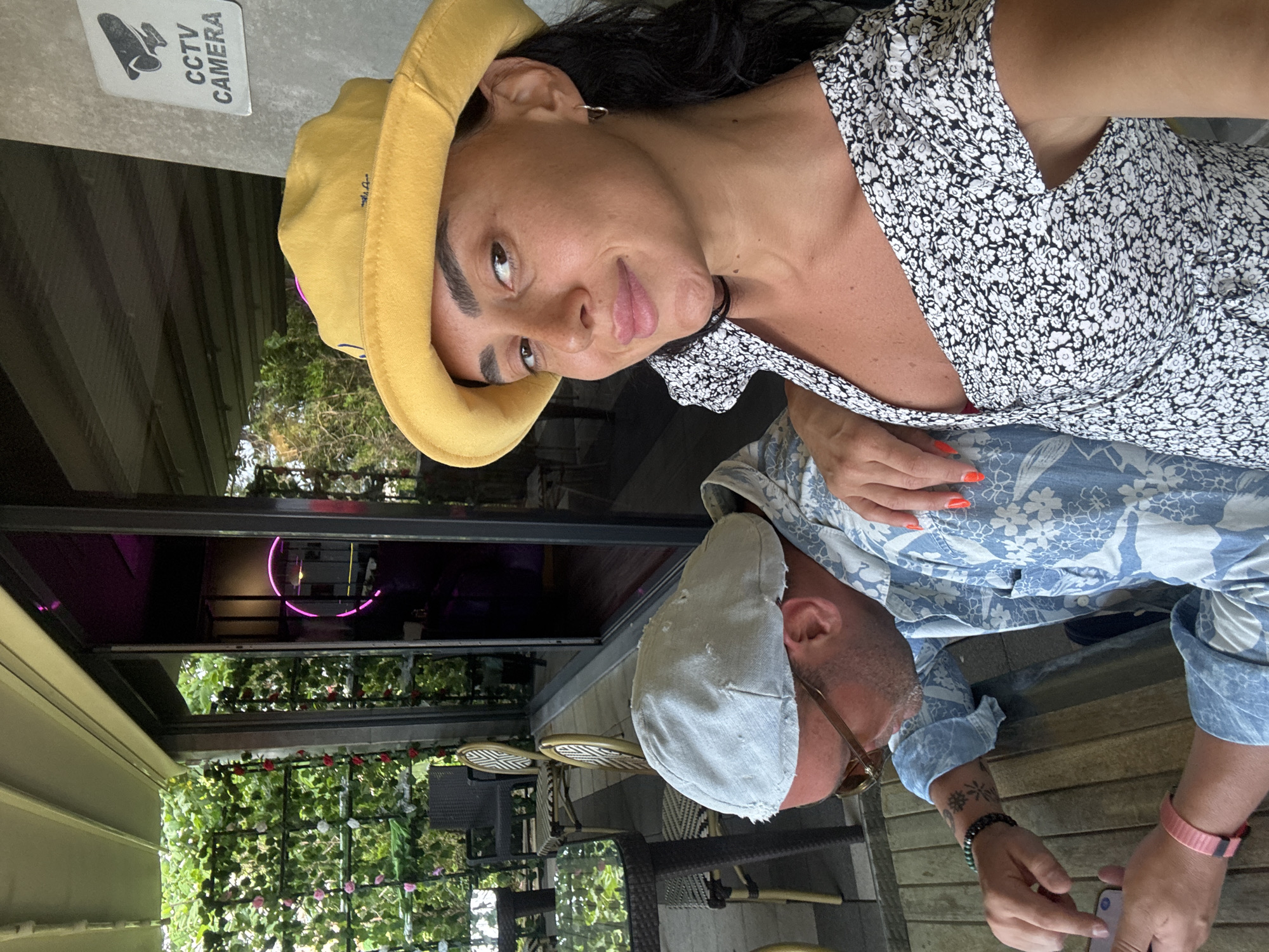 This is a selfie taken outdoors, likely on a covered porch or patio, at a cafe in Batumi, Georgia. The foreground features a woman with dark hair, wearing a yellow bucket hat and a black and white floral print dress. She is looking towards the camera and smiling. A man is visible to her left, wearing a light blue floral print shirt and a light-colored cap. He is looking down at a smartphone in his hands. The background shows a glimpse of an indoor space with neon lighting and an exterior area with green foliage and outdoor seating. A CCTV camera sign is visible on the wall in the upper right corner of the image.