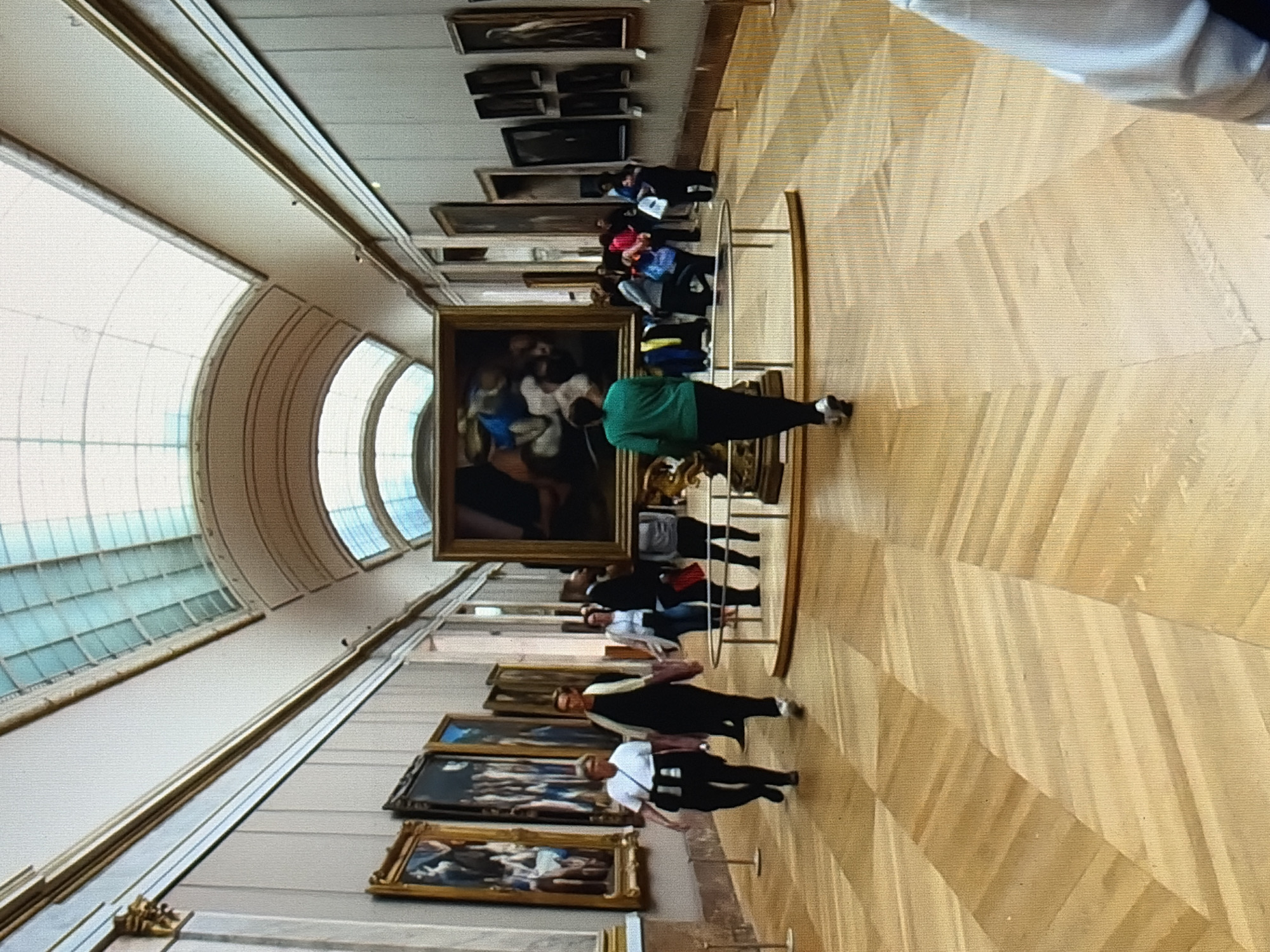 An indoor shot shows a long art gallery with a high, arched ceiling with a skylight. The walls are lined with paintings. Two women are walking on the left side of the gallery, and several other people are visible in the distance, some looking at artwork. The floor is made of polished wood with a geometric pattern. A large framed painting is displayed in the center of the gallery. The image is somewhat blurry and taken from a slightly low angle.