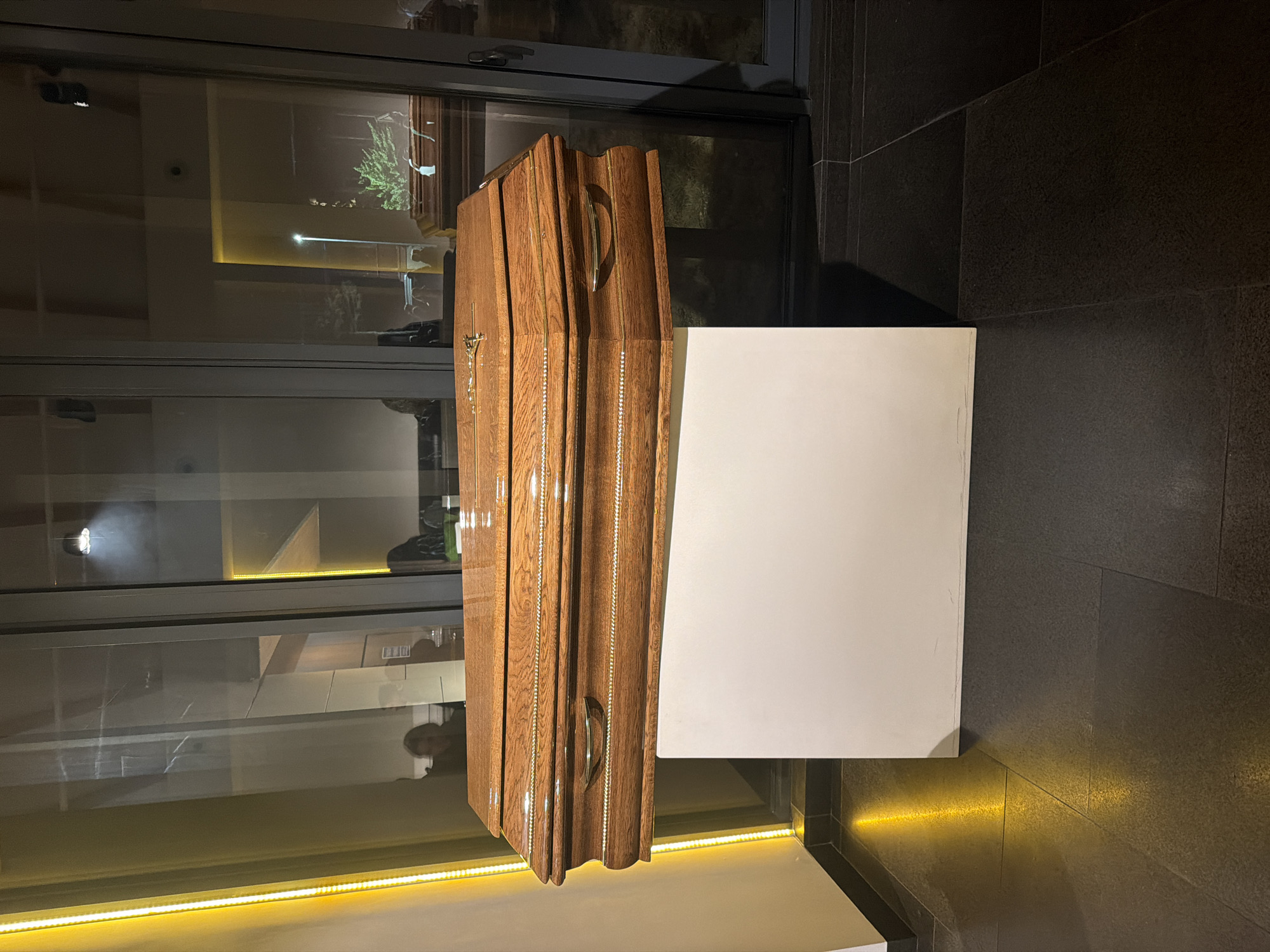 The image shows a polished wooden coffin standing upright on a white, square pedestal. The coffin is made of oak or a similar hardwood, with noticeable grain and a glossy finish. It has decorative handles and possibly a cross attached to its side. The pedestal is a plain white cube. The backdrop suggests an indoor setting, possibly a funeral home or memorial space, featuring dark tiled floors and a glass window reflecting interior elements. The location is stated as Unknown, Unknown. There are no people visible in the foreground, although a faint human form may be reflected in the window. The lighting in the scene is a mix of natural light and artificial illumination, creating highlights and shadows.