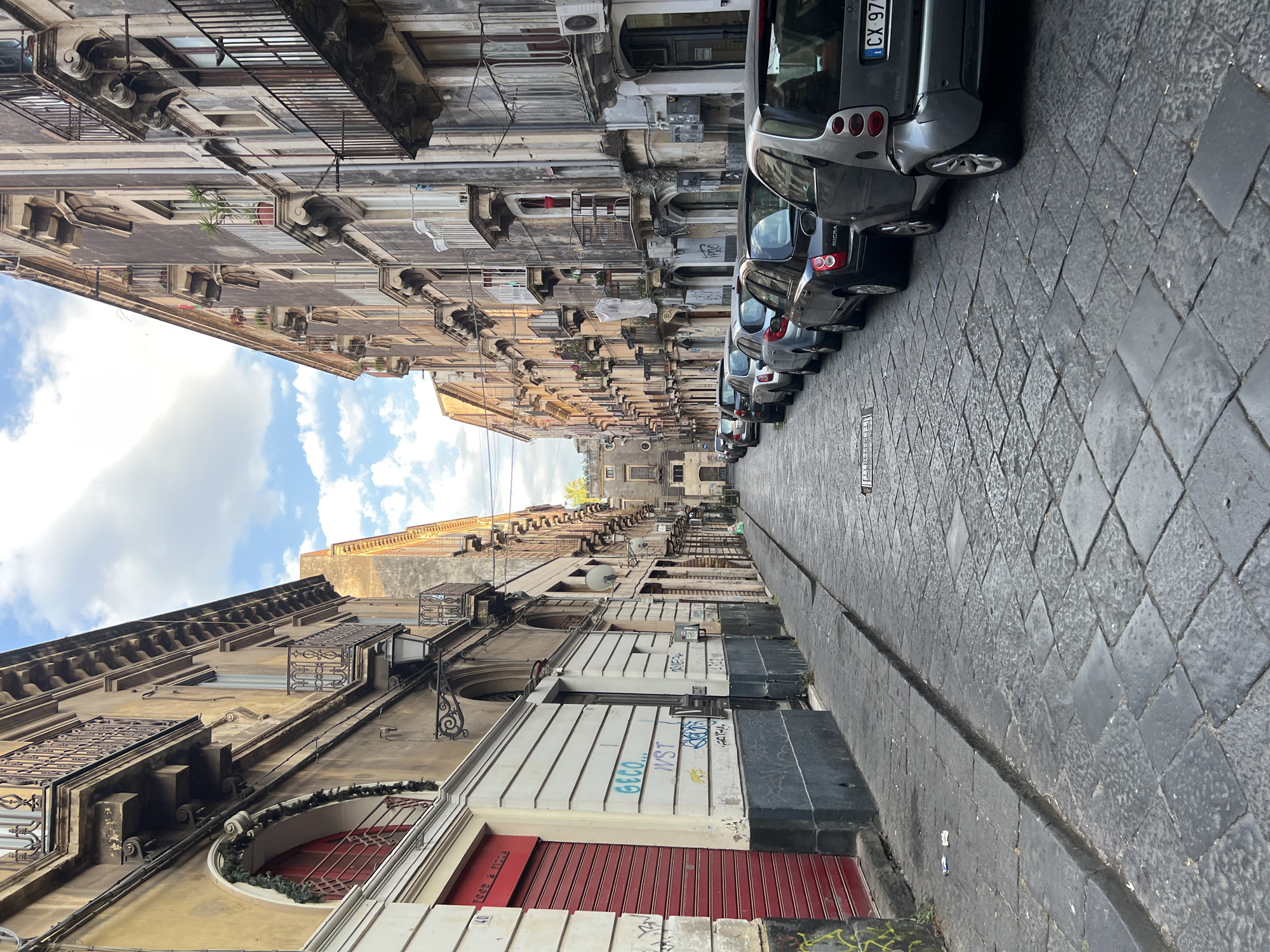 This is a vertical, eye-level shot of a narrow street in Catania, Italy. The street is lined with old, multi-story buildings with ornate balconies and architectural details. The buildings on the left side of the street are characterized by their weathered facades, some with visible graffiti. They feature arched windows, wrought-iron railings, and air conditioning units.
On the right side of the street, parked cars are visible, creating a visual boundary. The cars are mostly dark-colored and appear to be small to medium-sized. The street is paved with dark, diamond-shaped cobblestones, and a raised concrete curb runs alongside the parked cars.
The sky is visible in the upper left quadrant of the image, with some clouds and sunlight suggesting it is daytime, likely morning or afternoon. The overall impression of the scene is one of an older, urban environment, possibly a historic district, with a mix of residential and commercial buildings. There is no visible activity of people in the frame.
Some visible text includes a license plate on one of the cars with "CX 970..." and some graffiti on the building facades, including "GECO...", "IVST", and other illegible markings. Another visible text on a red shutter reads "FOOD & PIZZA". The narrowness of the street and the stacked architecture create a sense of enclosure.