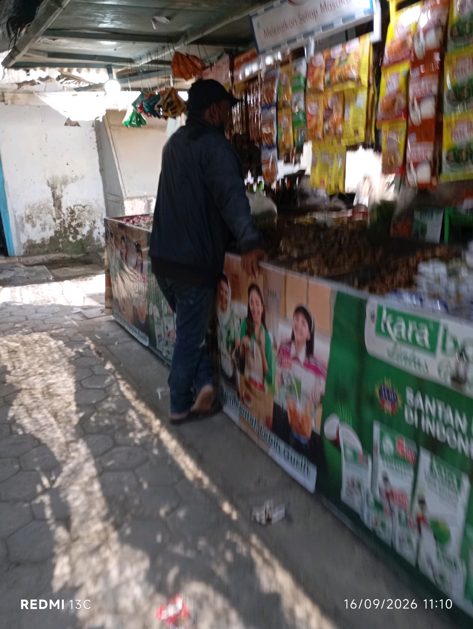 A middle-aged man wearing a dark jacket and a black baseball cap stands near a roadside stall in Wuluhan, Indonesia. He is leaning on the stall, which is adorned with colorful advertisements for various food products. The stall appears to be constructed with a wooden frame and covered with a corrugated metal roof. Behind the man, the stall is filled with packaged goods, predominantly snacks and food items. The ground in front of the stall is paved with hexagonal tiles and illuminated by dappled sunlight, casting long shadows. To the left of the stall, a weathered white wall with some discoloration is visible. The overall scene depicts a typical street-side market environment.