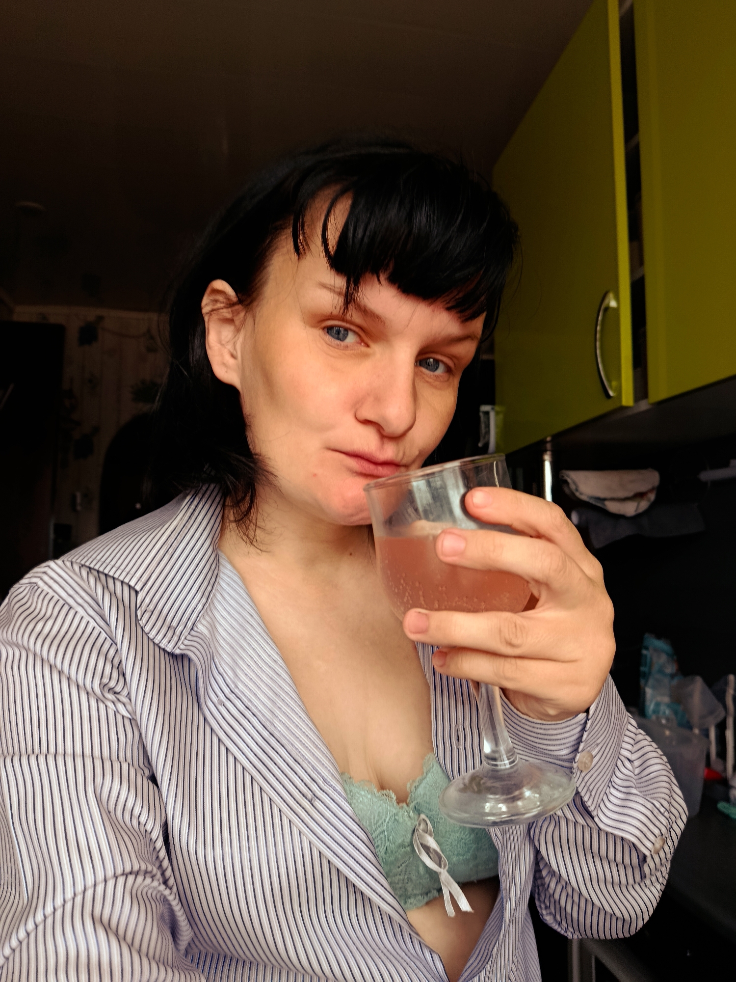An indoor, eye-level portrait captures a woman with dark hair, bangs, and striking blue eyes, looking directly at the viewer. She is wearing an unbuttoned, light blue and white striped long-sleeved shirt, revealing a light green lace bra underneath. Her left hand holds a clear stemmed glass filled with a pinkish, possibly effervescent beverage, bringing it to her lips as if taking a sip or about to.
The background suggests a domestic setting, likely a kitchen, in Фоки, Russia. To the woman's right, a vibrant lime-green kitchen cabinet is prominent, contrasting with a darker countertop below. The area behind her on the left is darker, revealing hints of a wall with a pattern. The lighting appears to be natural, possibly from a window, illuminating her face and the front of her body. Her expression is calm and direct, with a subtle hint of a smirk. There is no visible text in the image.
