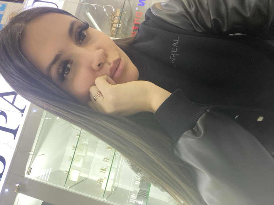 A female individual is depicted in a close-up, self-portrait photograph taken inside a retail store in Chaikovsky, Russia. She has long, dark hair and prominent eyelashes, with her left hand positioned against her cheek. Three thin, golden-colored rings are visible on her left ring finger. She is wearing a black long-sleeved garment, possibly a jacket or sweatshirt, featuring contrasting shiny black sleeves and ribbed cuffs. The word "UNREAL" is embroidered in black on the chest of her garment. In the background, a brightly illuminated glass display cabinet is visible, stocked with multiple white and clear bottles, consistent with perfume products, each bearing a "LE PARFUM" label and a gold-colored cap. Above this display, a partial sign displaying large capital letters, including "PARF" or "SPAR", is present, along with draped white string lights. The overall scene indicates an indoor commercial setting, specifically a perfume or beauty retail environment.