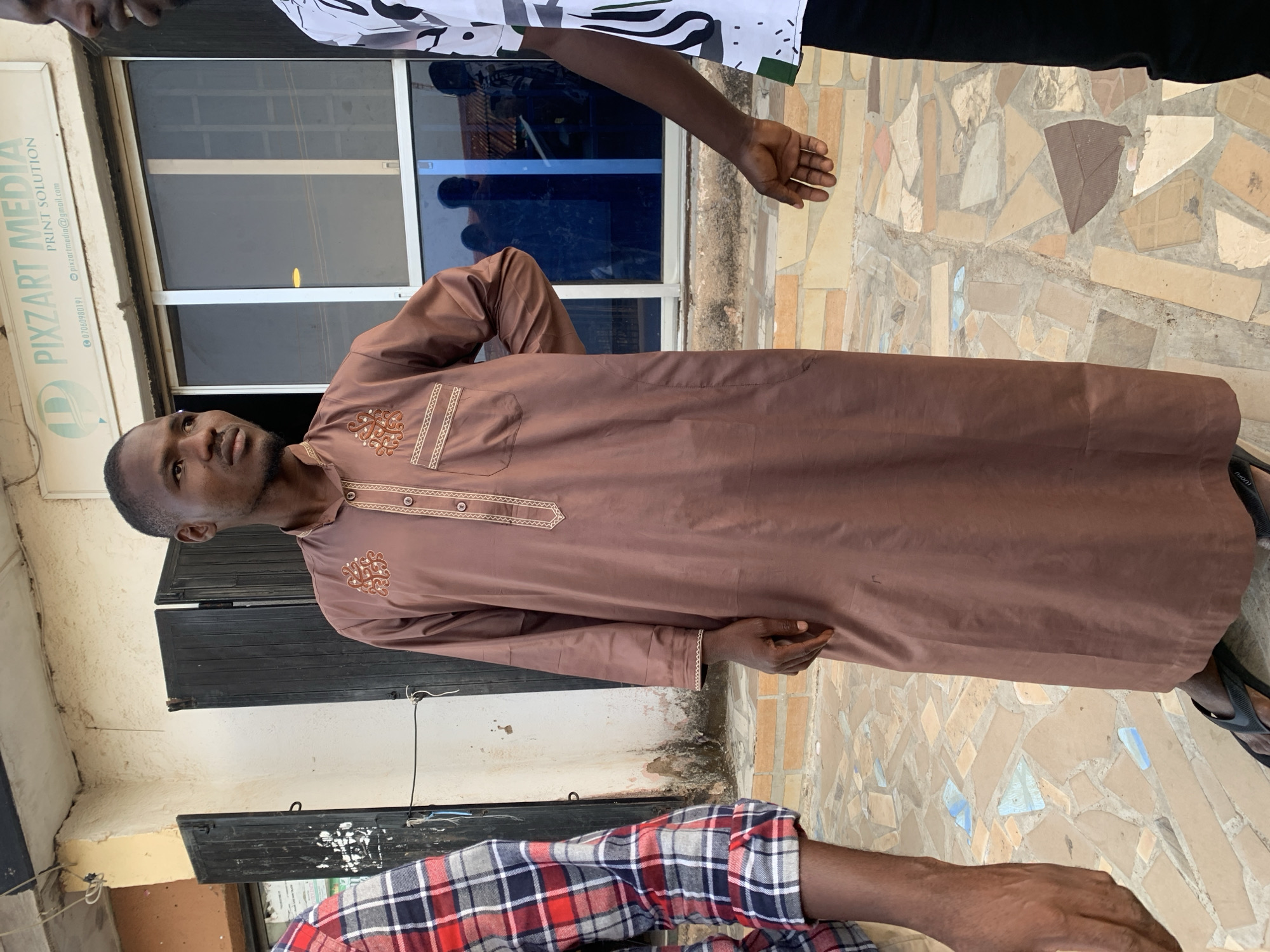 A man wearing a brown traditional robe with embroidered details stands outside a building with "PIXZART MEDIA PRINT SOLUTION" signage, presumably in Jos, Nigeria. The building has windows, some reflecting silhouettes. The ground is paved with light-colored tessellated tiles. Parts of two other individuals are visible: one wearing a white and black patterned shirt and the other in a red and blue plaid shirt.