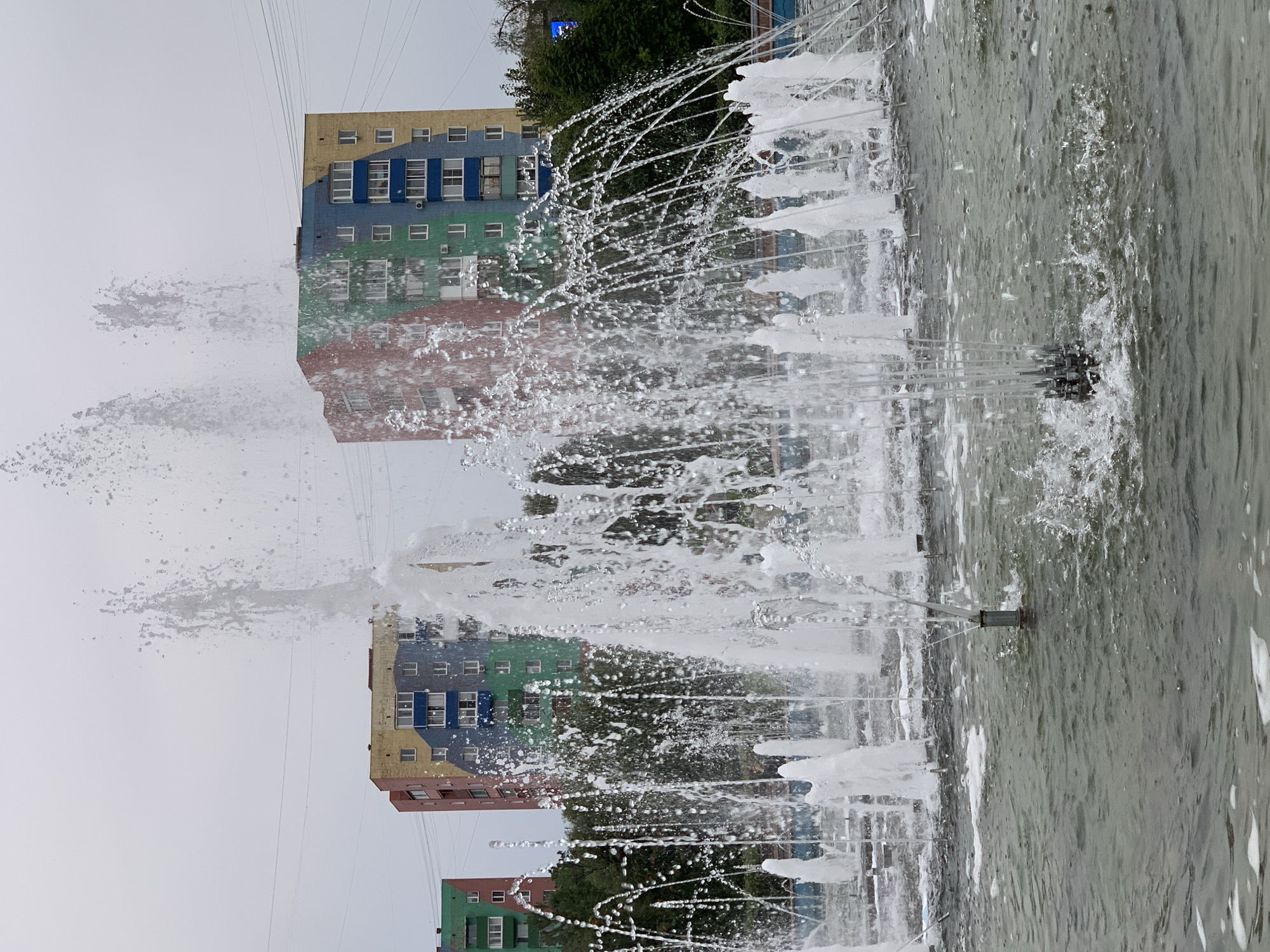 In Prokopyevsk, Russia, a fountain is active in front of three colorful apartment buildings. The buildings are painted in blocks of yellow, blue, red, and green. The fountain sprays water in various streams and arcs, with some water hitting the surface of the pool and creating splashes. The sky is overcast. The image is taken at eye-level, looking towards the buildings and the fountain. No people are visible in the image.