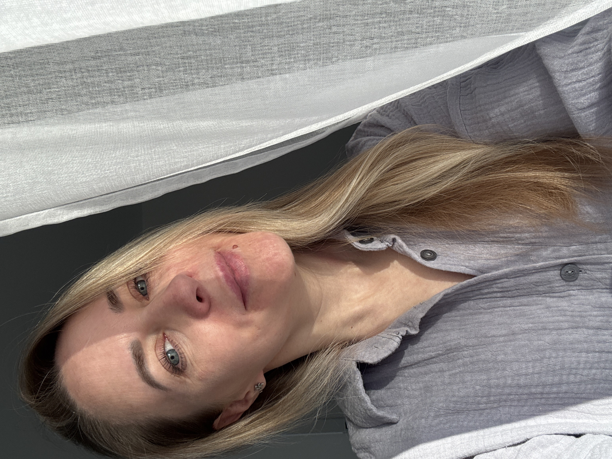 A woman with blonde hair and blue eyes is lying down, likely indoors, based on the soft, diffused lighting. She is wearing a gray collared shirt. A sheer white curtain is partially visible above her. The location in the city of Chaikovsky, Russia cannot be determined from the image.
