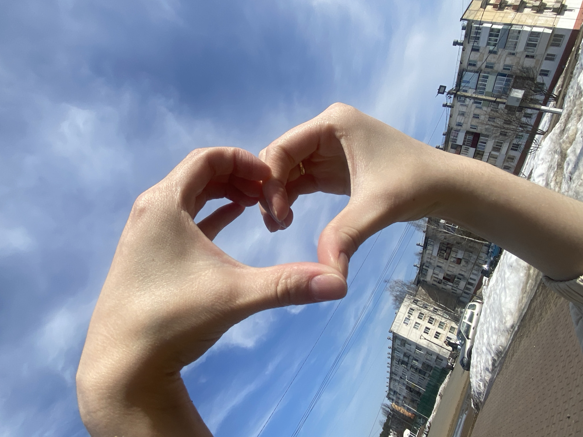 Here is a description of the image:
The image shows two fair-skinned hands creating a heart shape against a bright blue sky with streaky white clouds. A gold ring is visible on the left hand. The background includes apartment buildings, bare trees, patches of snow, a road, and power lines, suggesting an urban environment. Based on these characteristics the location can be identified as Chaikovsky, Russia.